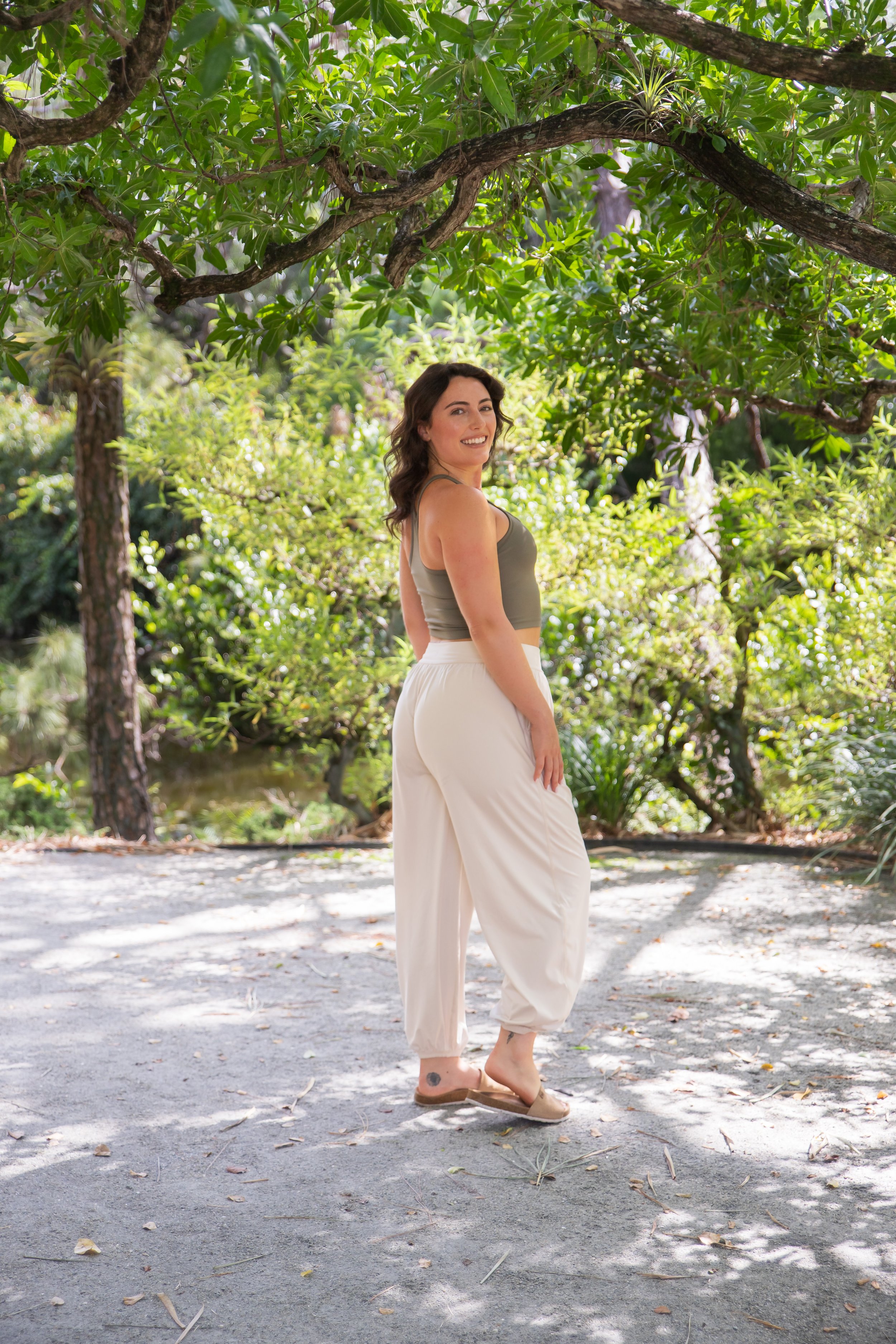 A woman with dark wavy hair smiling and standing outdoors on a dirt path, surrounded by green trees and sunlight filtering through the leaves.