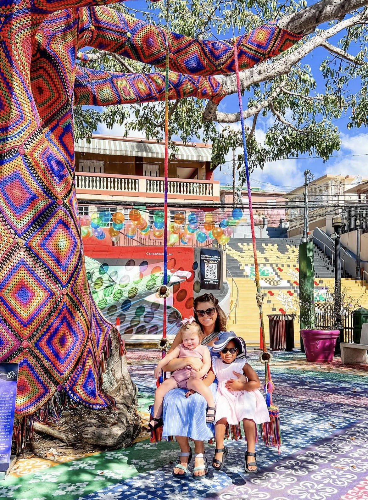 Just Another Wagner Adventure | Two young girls and a woman sitting on a swing under a large tree, with colorful decorations and buildings in the background. Yauco in Puerto Rico.