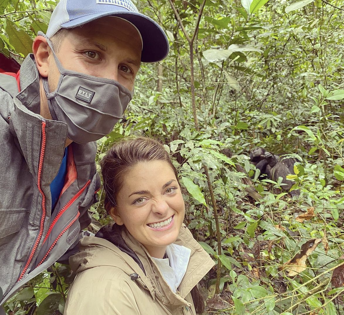 Just Another Wagner Adventure | A man and woman taking a selfie in a dense, green forest with chimpanzee in background. Kibale National Park in Uganda.