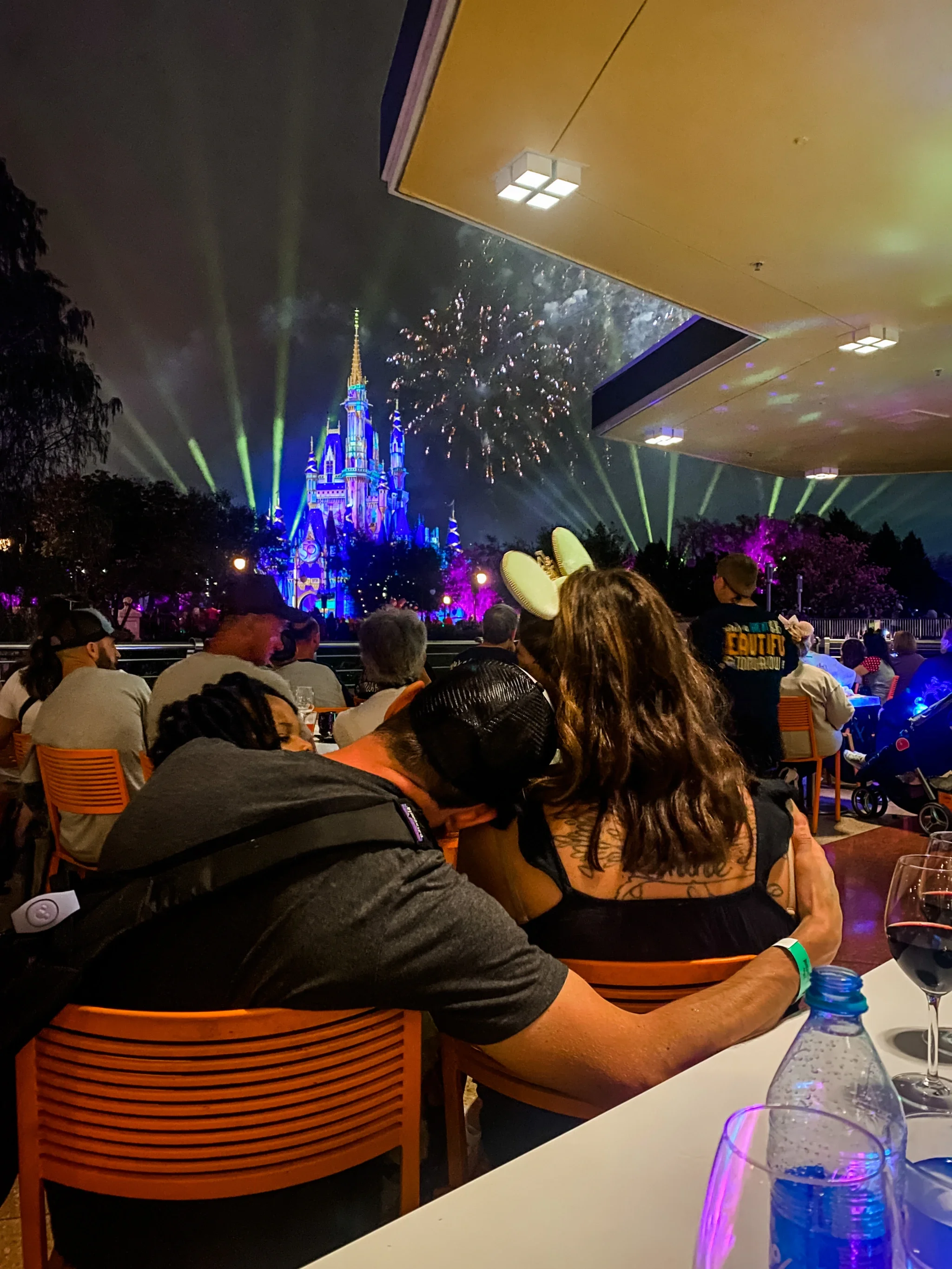Just Another Wagner Adventure | People sitting at an outdoor table watching fireworks and a colorful castle light show at night. Magic Kingdom, Walt Disney World, Florida.