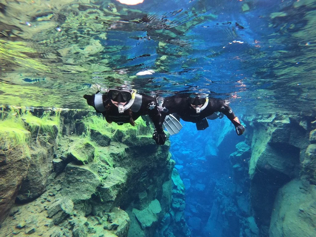 Just Another Wagner Adventure | Two people in wetsuits scuba diving in an underwater canyon or crevice with rocky, moss-covered walls. Thingvelir National Park in Iceland.