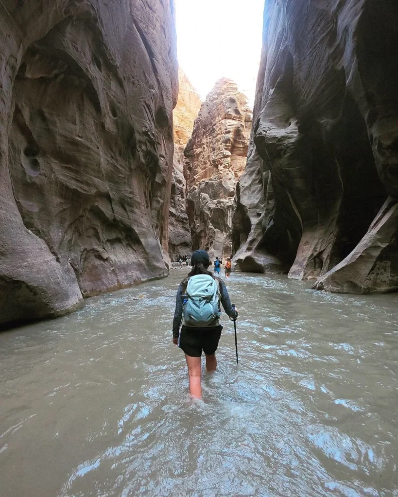 Just Another Wagner Adventure | A person hiking through a narrow canyon with steep, high walls, crossing a shallow water stream. The Narrows in Zion National Park, Utah.