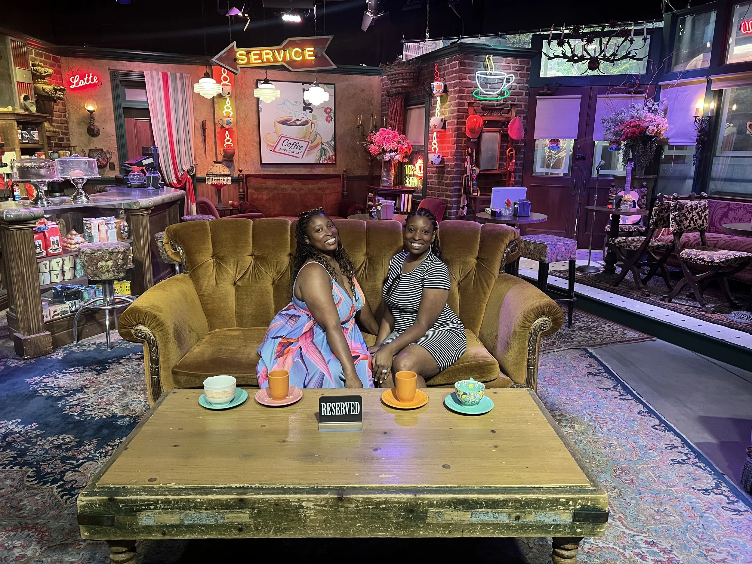 Just Another Wagner Adventure | Two women sitting on a yellow vintage sofa in a cozy, decorated cafe or coffee shop with vibrant neon signs, floral arrangements, and a wooden table with cups and a reserved sign.