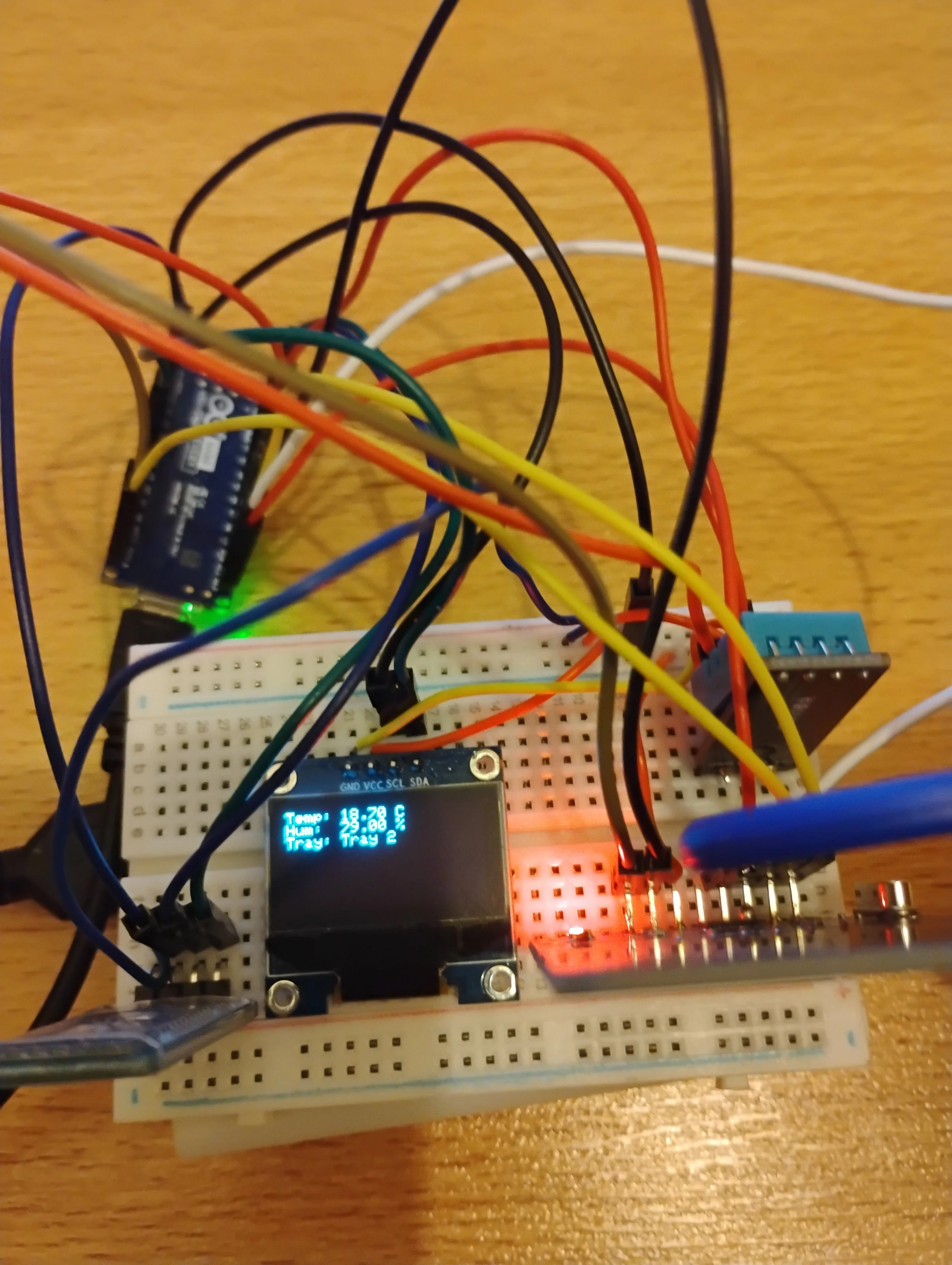 A breadboard with an Arduino microcontroller, a small OLED display showing temperature and humidity, multiple connected wires, a temperature sensor, and several other electronic components.