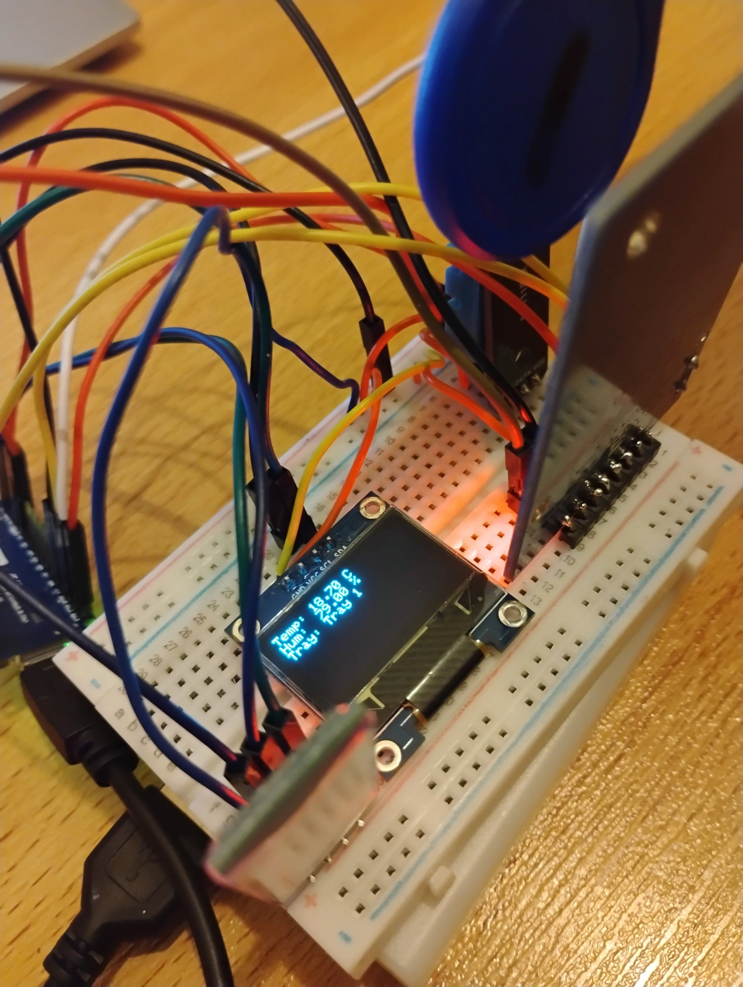 Electronic circuit with wires, a breadboard, a microcontroller, a display showing temperature, humidity, and pressure readings, and a component with a blue circular top.