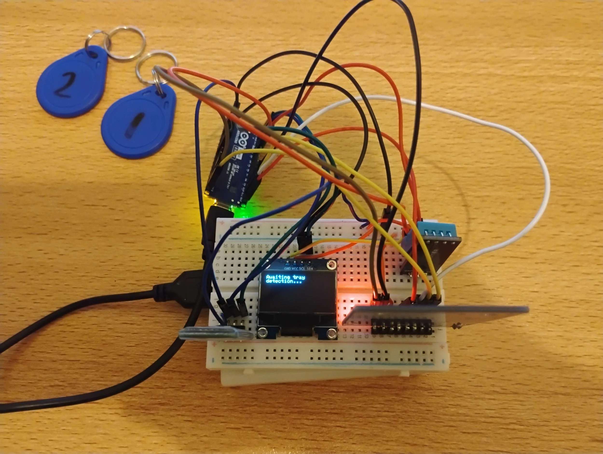 A breadboard with electronic components, including a small display showing 'Waiting for detection...' and various wired sensors, connected to a microcontroller, with two blue keychains with numbers '1' and '2' on it, placed on a wooden surface.