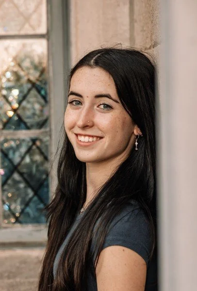 A woman with dark wavy hair, wearing a red dress and hoop earrings, smiling and posing for a portrait.
