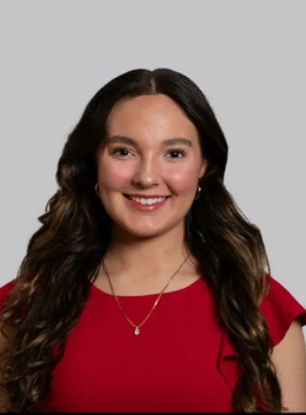 Young woman with long black curly hair smiling at the camera, outdoors with greenery in the background.