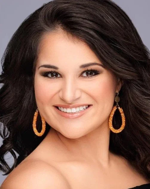 A woman with long blonde hair, blue eyes, and makeup, smiling and wearing large sparkly earrings and a black top.