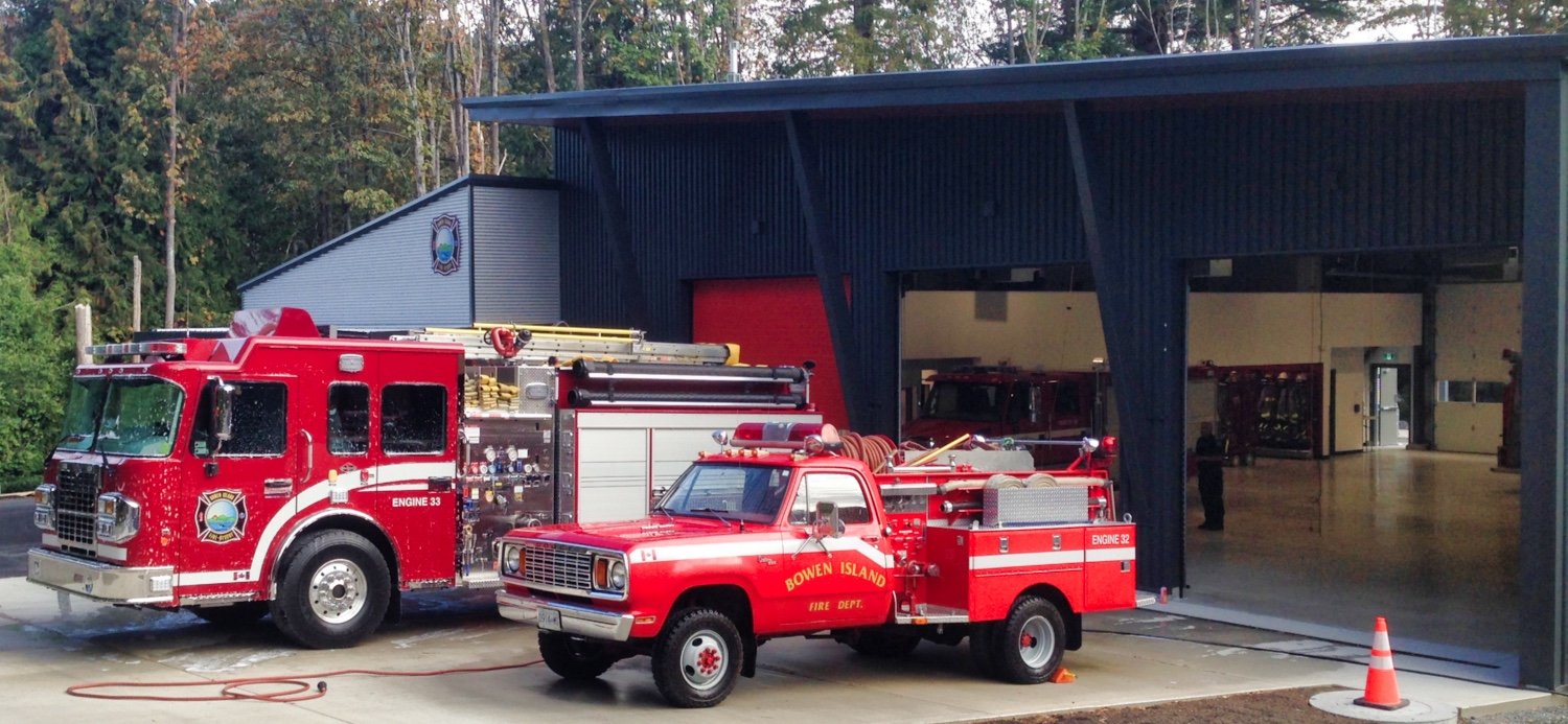 Two fire trucks parked outside a fire station garage. One large red fire truck with a ladder and equipment, and a smaller red fire department pickup truck labeled 'Bowen Island Fire Dept.'