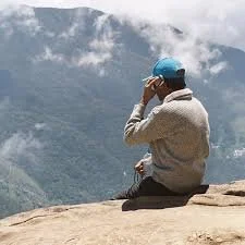 Person sitting on rocky ledge wearing a blue helmet, overlooking mountainous landscape