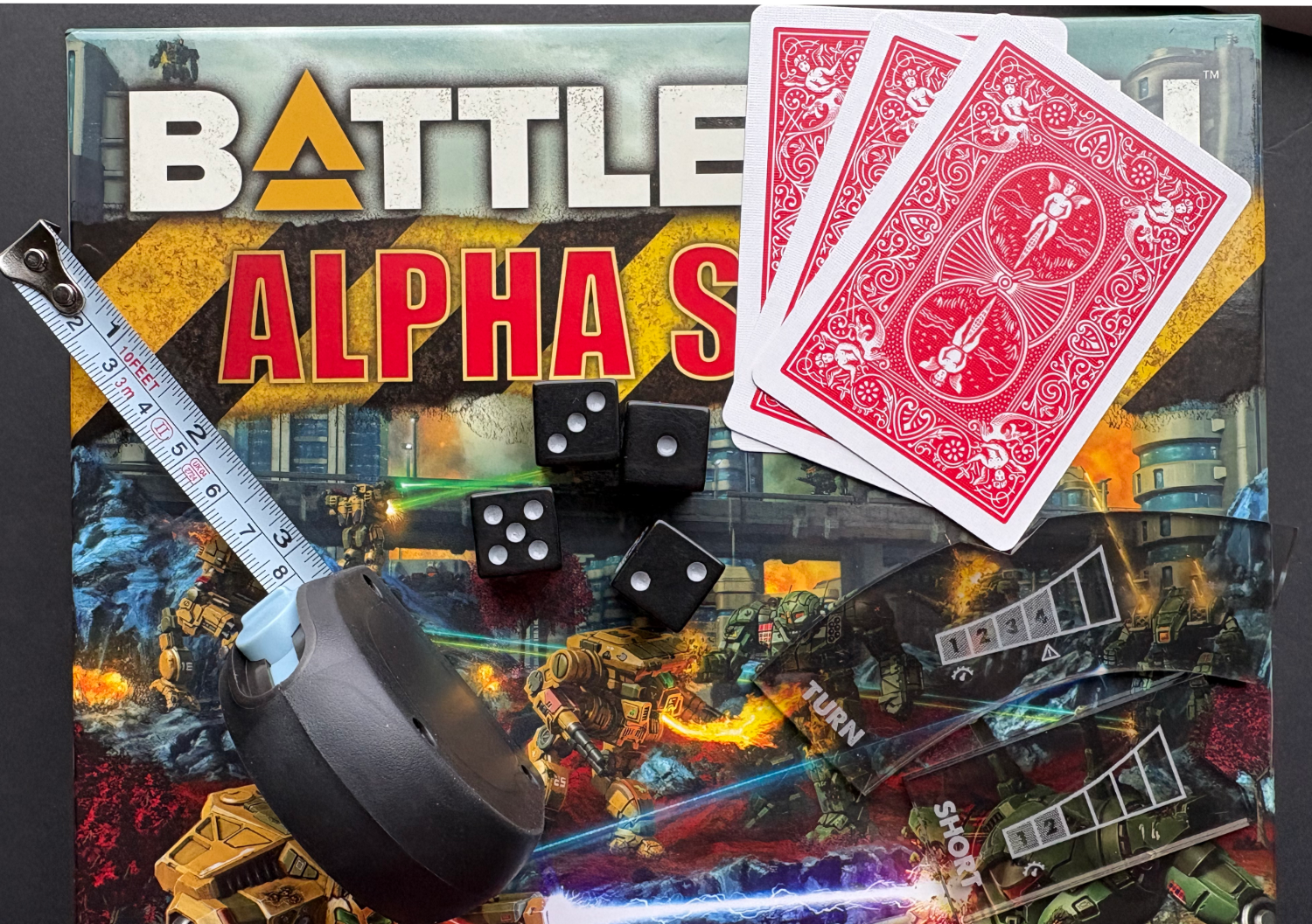A tape measure, black dice, Gaslands movement templates, and a deck of playing cards arranged on a Battletech Alpha Strike box