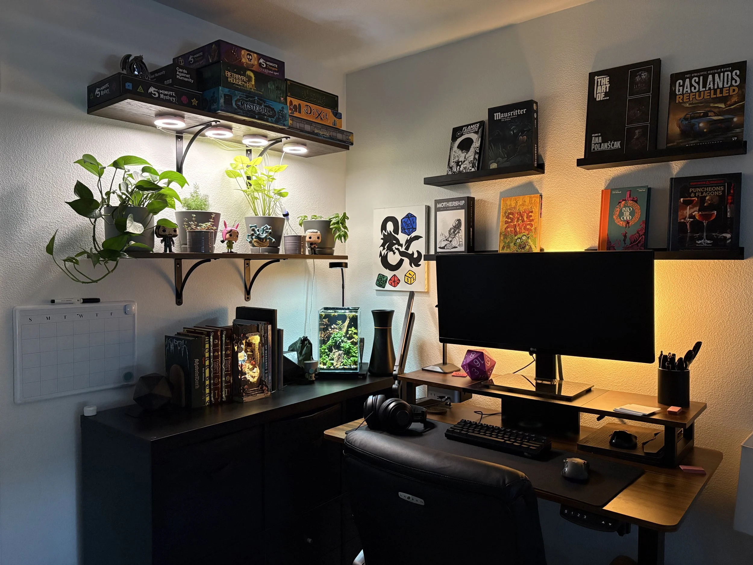 A cluttered home office with two shelves filled with board games, books, and plants, a desk with a computer monitor, keyboard, mouse, and headphones, a small fish tank, and various small decorative items.