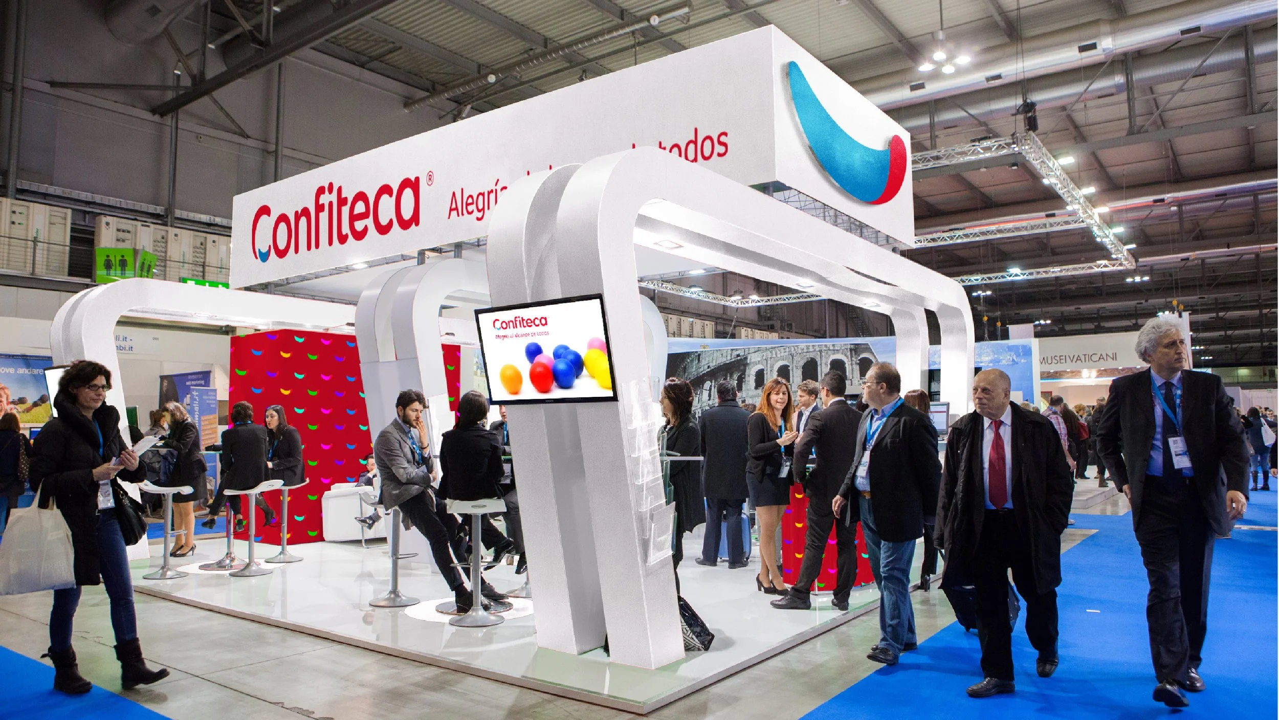 Stand de Confiteca en feria internacional, con diseño moderno en blanco, pantalla digital y eslogan “Alegría al alcance de todos”. Visitantes interactúan en un entorno corporativo y dinámico, destacando la presencia de marca en eventos de negocios.