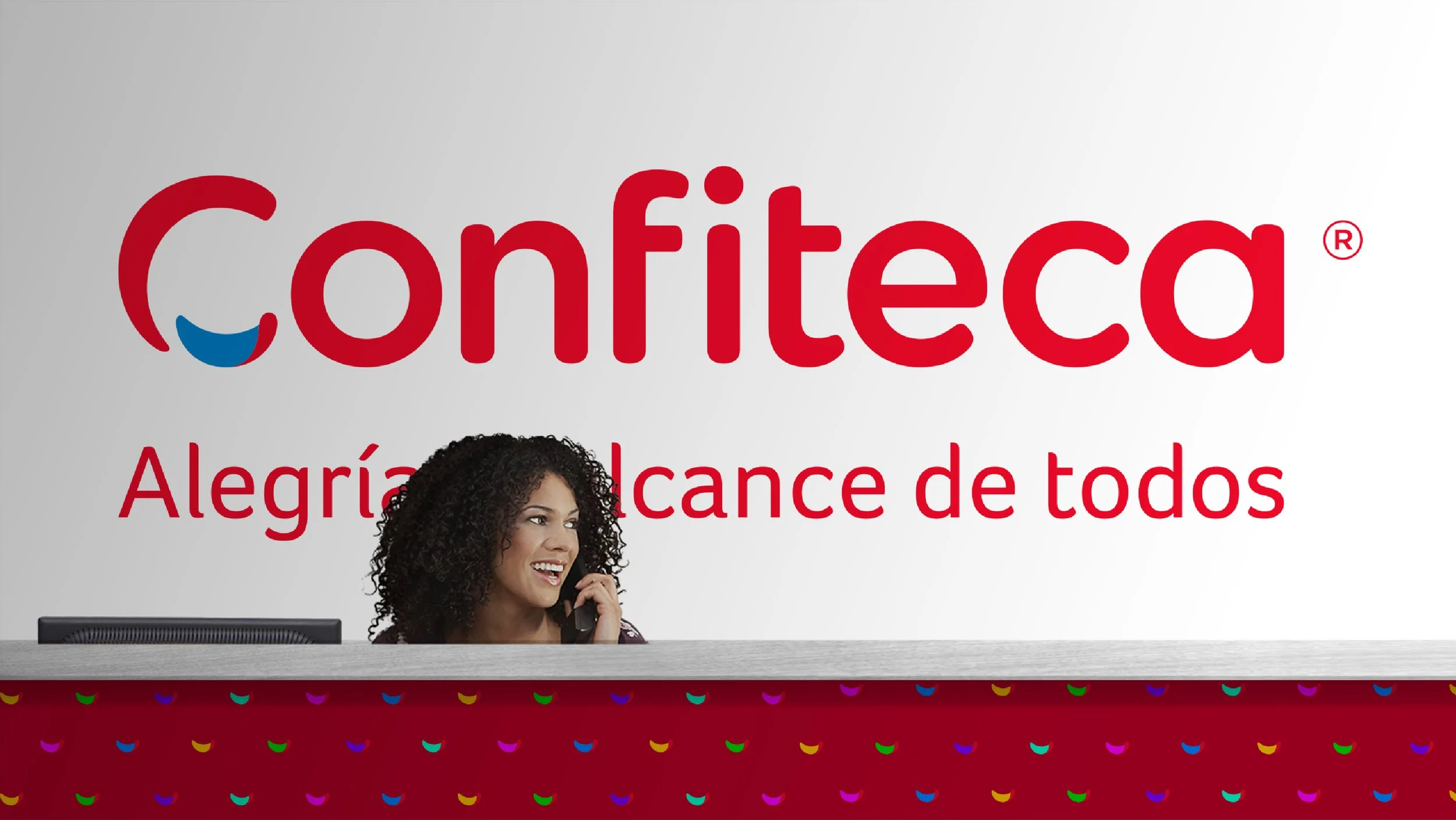 Recepción corporativa de Confiteca con logotipo rojo y eslogan “Alegría al alcance de todos” en fondo blanco. Mujer sonriente con cabello rizado atiende el teléfono tras un mostrador decorado con íconos coloridos, transmitiendo calidez, cercanía y pr