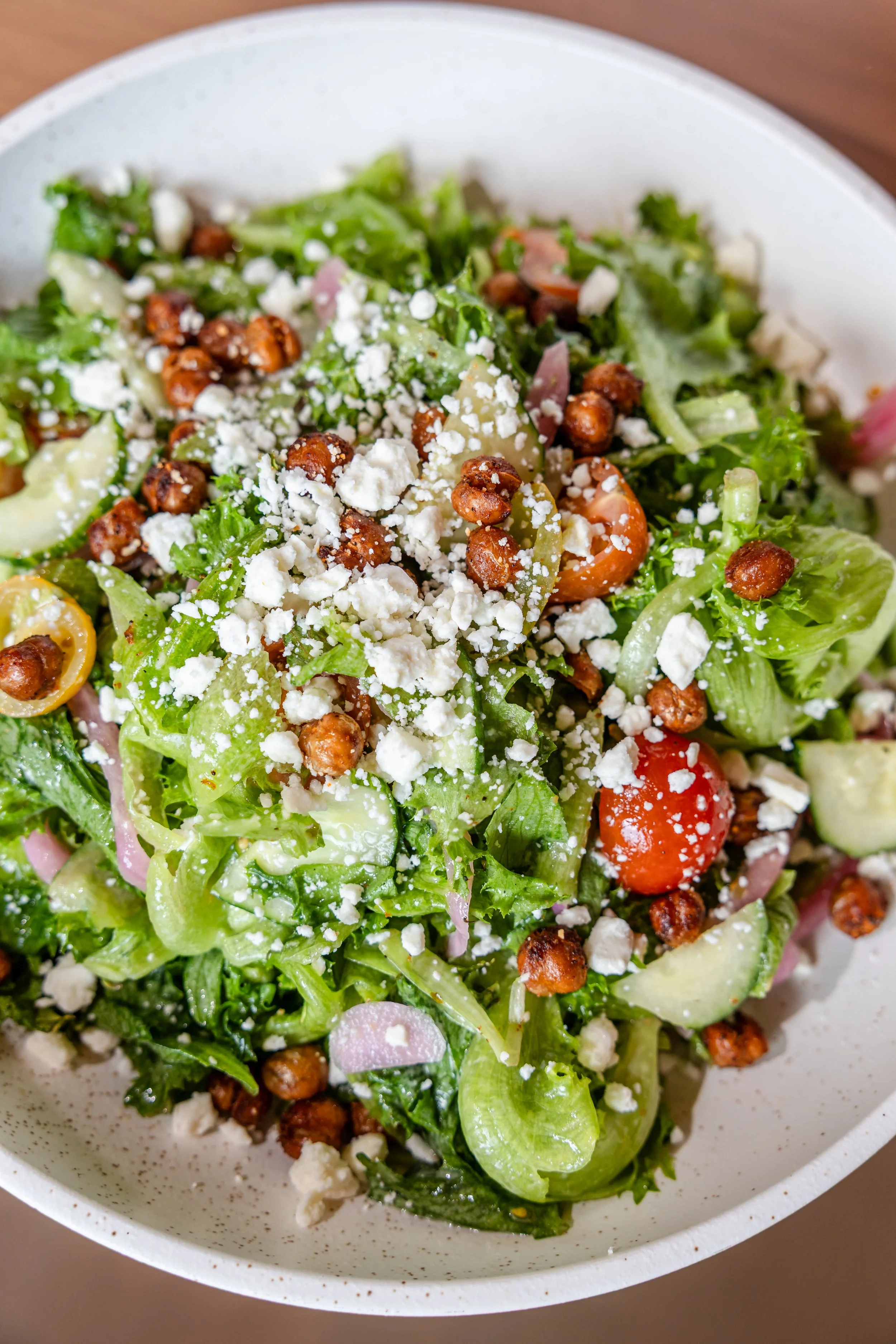 Close-up of a fresh salad with greens, cherry tomatoes, crumbled cheese, and toasted chickpeas in a white bowl.
