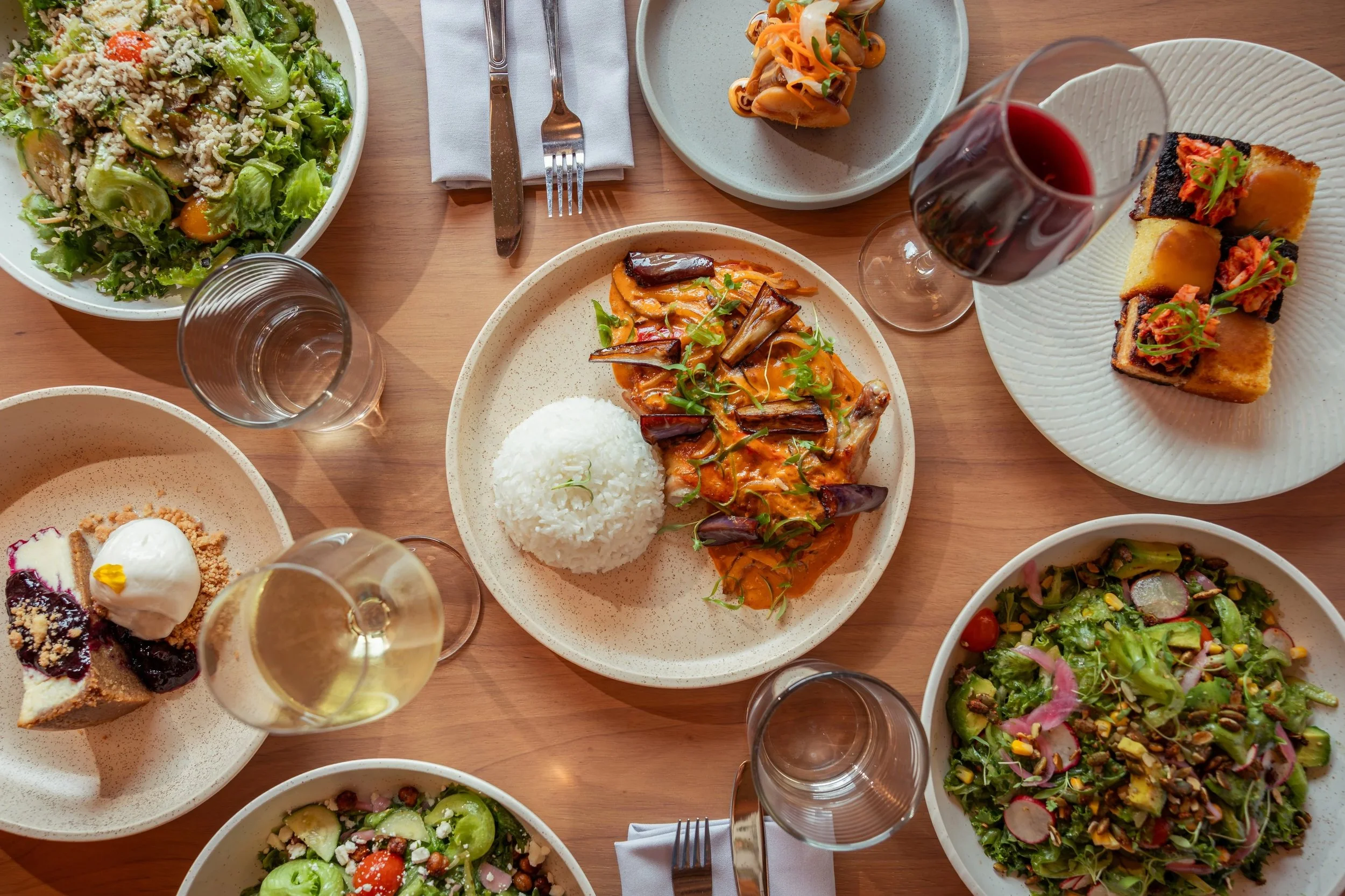 An overhead view of several plates and glasses on a wooden table, featuring salads, bread, a baked dish with rice, desserts, and glasses of red and white wine.