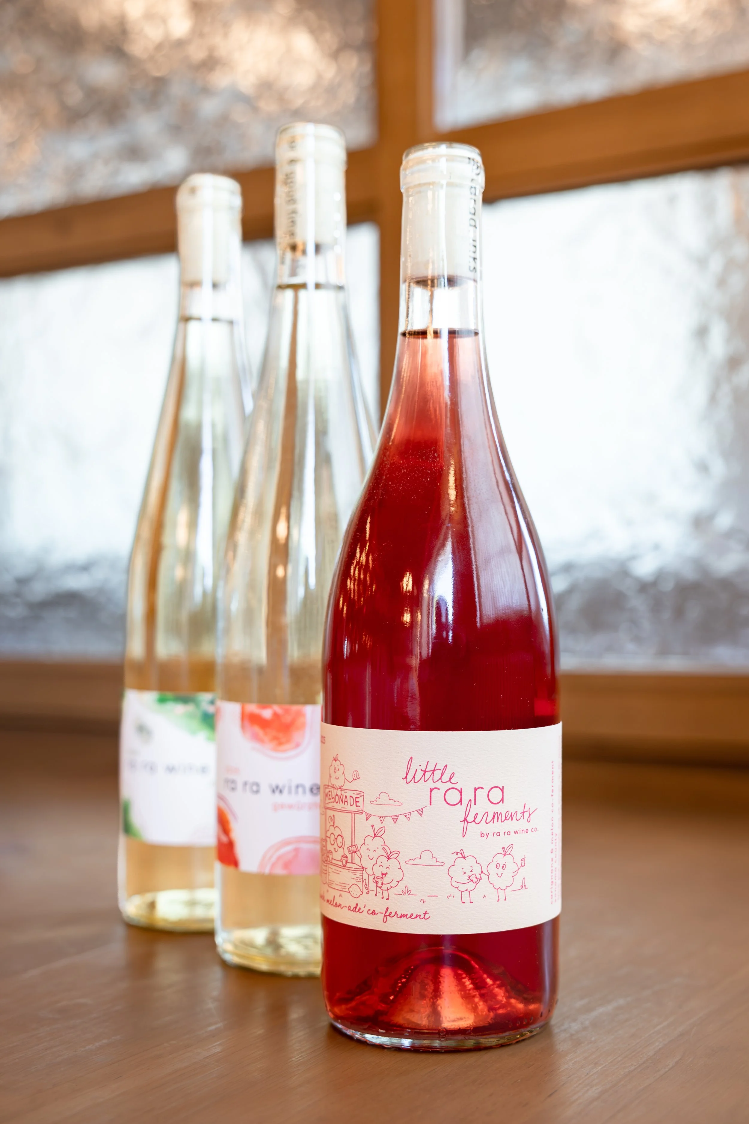Three bottles of fermented fruit beverage on a wooden table, with one bottle labeled 'Little Rara Ferments' in the foreground and two more bottles with colorful labels in the background.