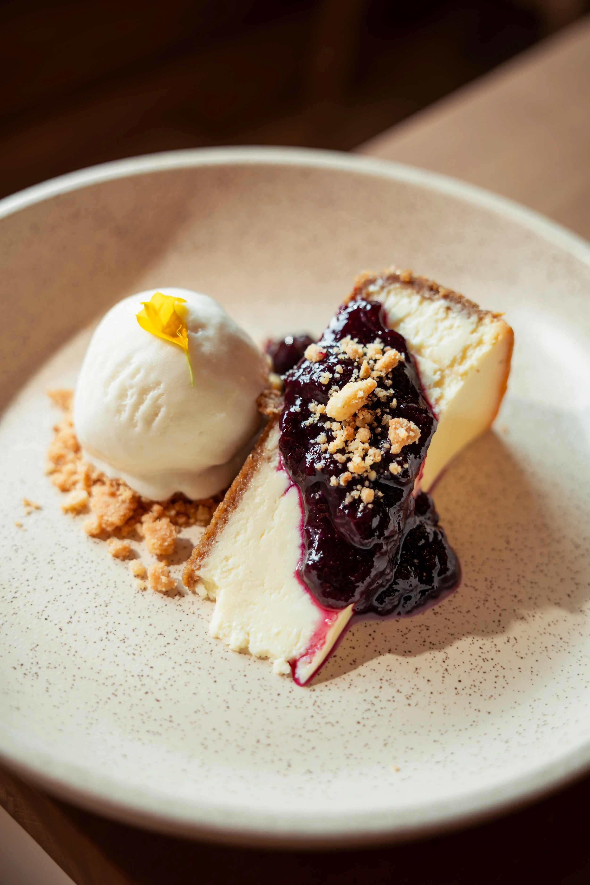 Slice of cheesecake with berry topping, garnished with a yellow flower petal, on a speckled beige plate
