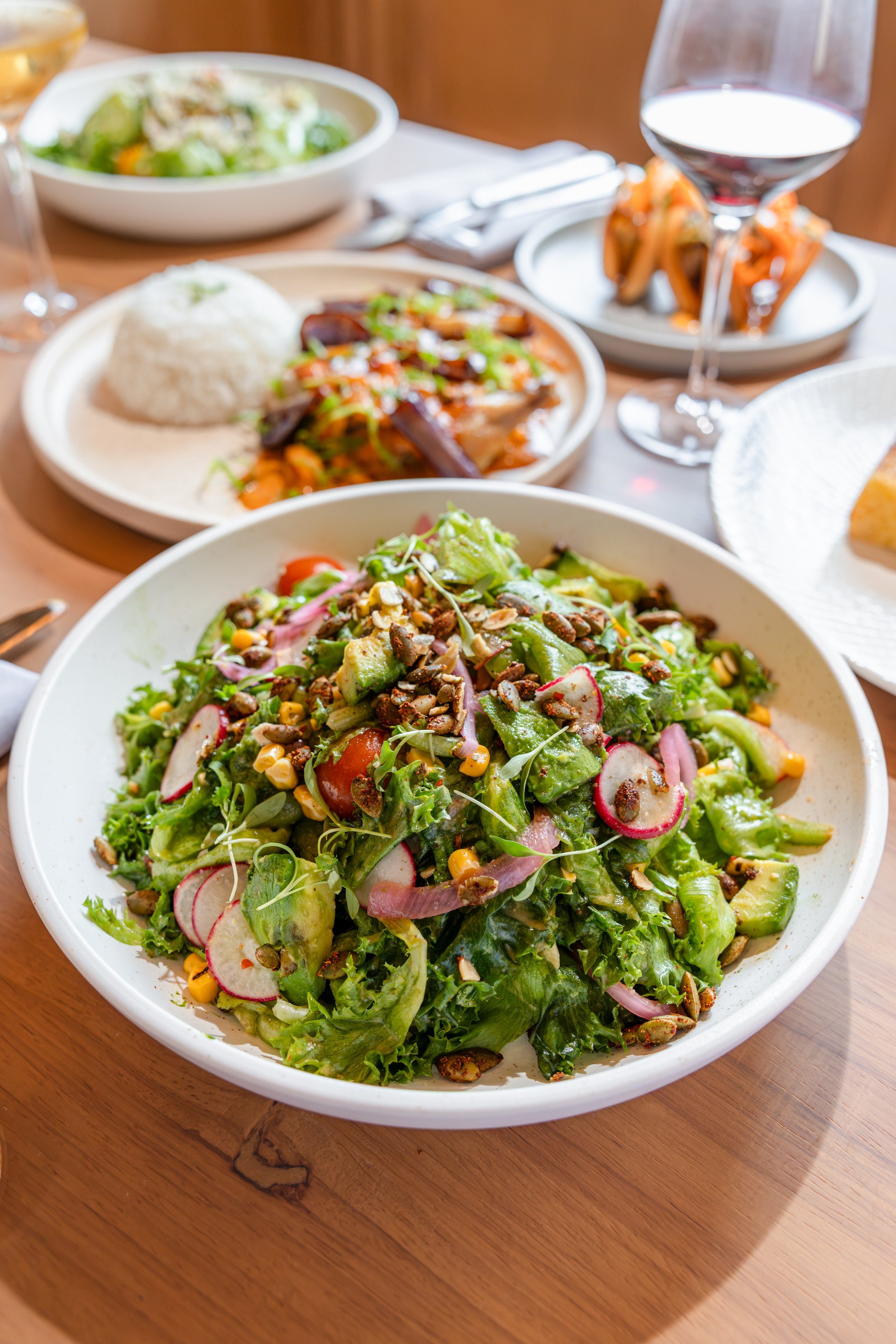 A bowl of salad with mixed greens, radishes, cherry tomatoes, avocado, corn, and seeds on top, placed on a wooden table.