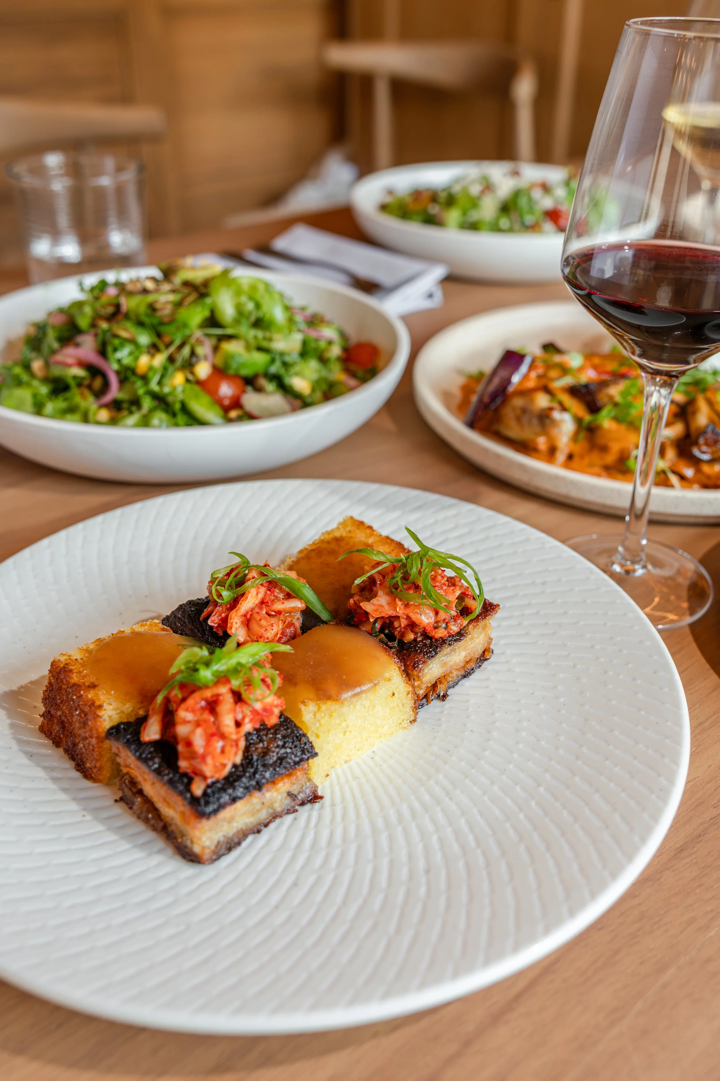A white plate with a dessert topped with kimchi, served on a white textured plate. In the background, there are salads in white bowls, a glass of red wine, a clear glass of water, and a wooden table setting.