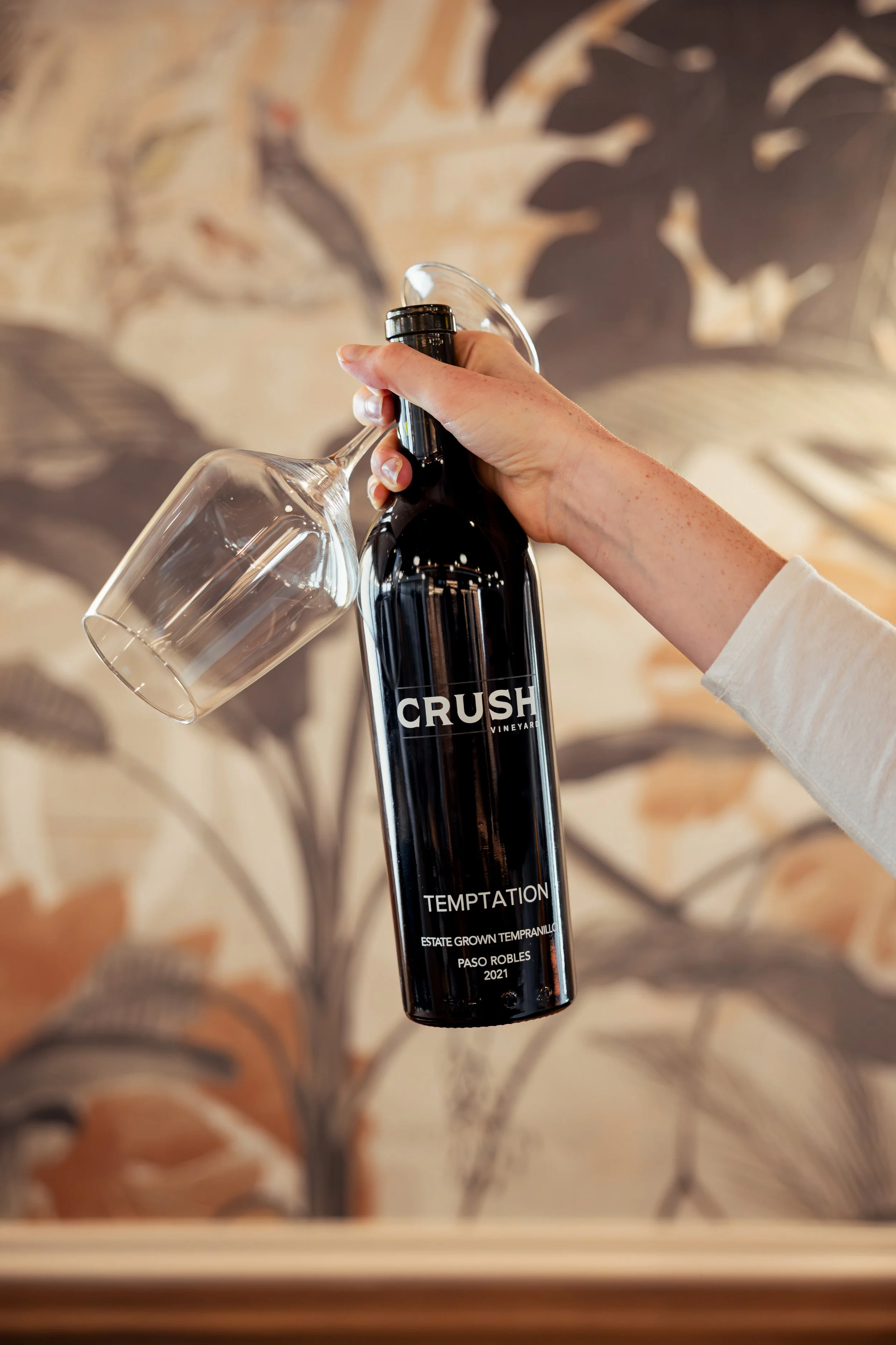 A hand holding a glass of white wine and a bottle of Crush Vineyard Temptation Cabernet Sauvignon.