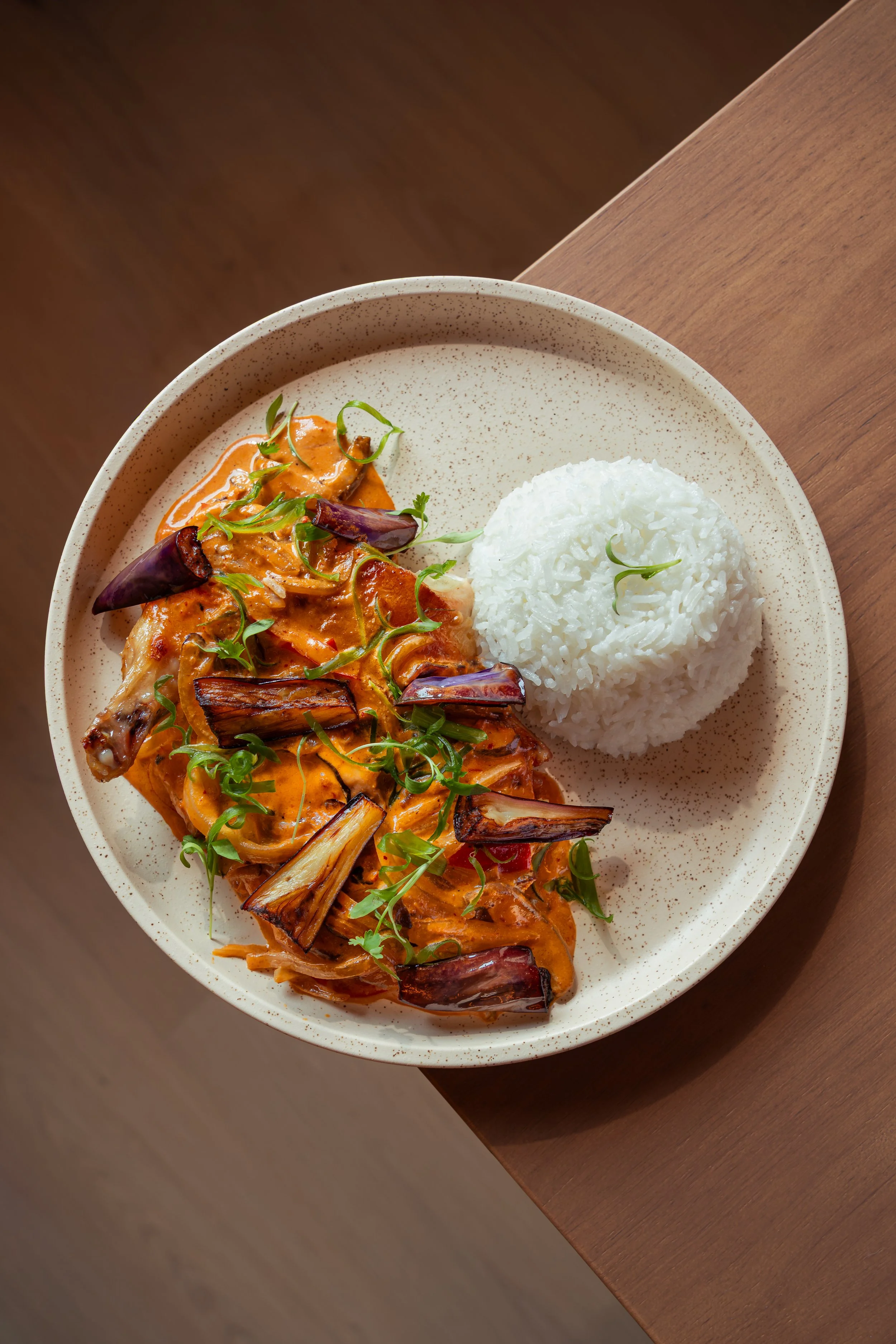 Plate of grilled chicken with reddish-orange sauce, garnished with green herbs, served with a side of white rice, on a speckled ceramic plate.