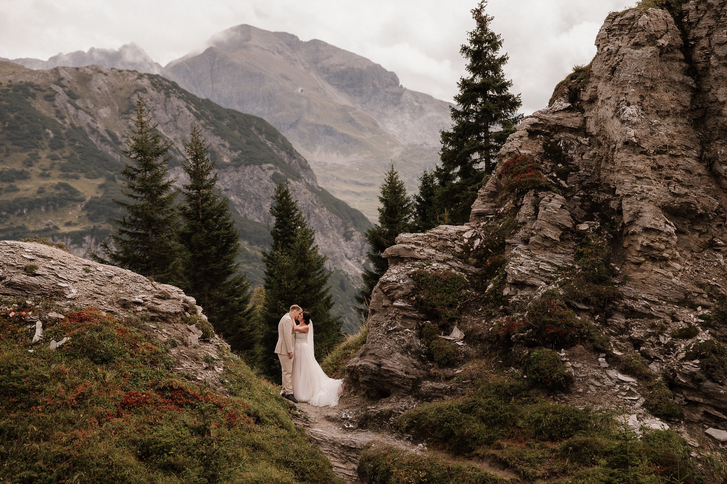 Ein Brautpaar, das sich liebevoll küsst, in einer Berglandschaft mit hohen Felsen, Tannenbäumen und bewölktem Himmel.