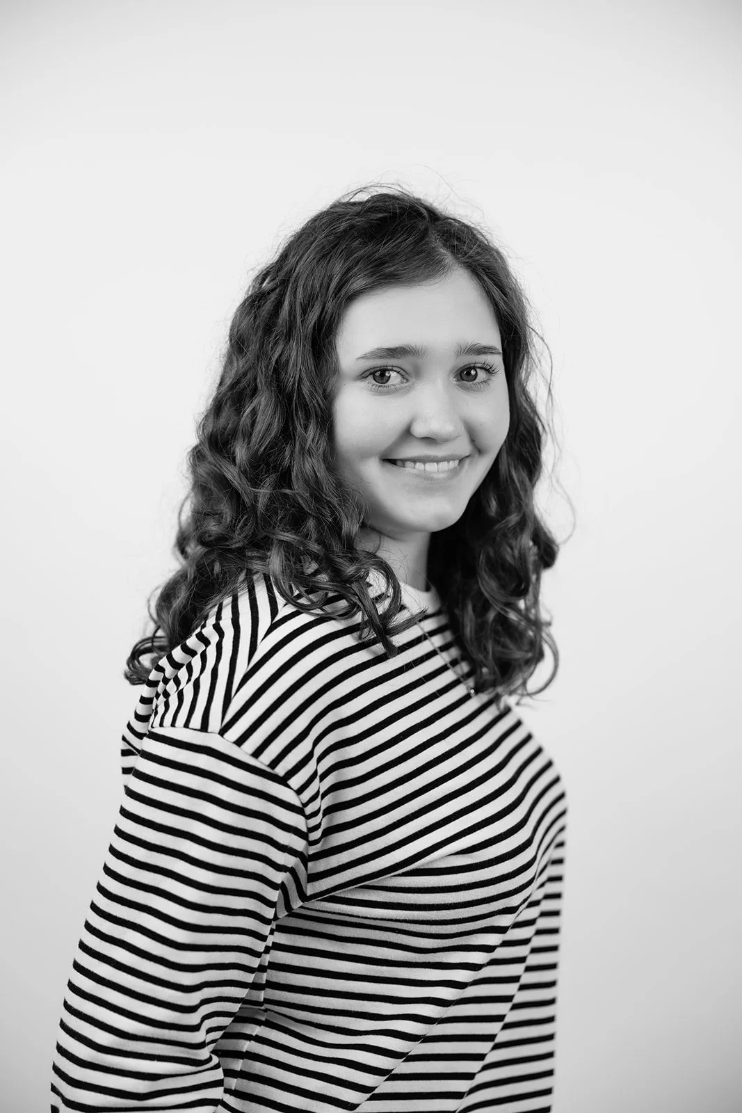 Schwarz-Weiß-Foto einer lachenden jungen Frau mit lockigem Haar, trägt ein gestreiftes Shirt, vor hellem Hintergrund.