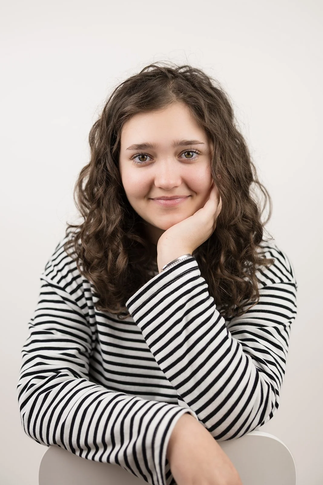 Porträt einer jungen Frau mit braunen, lockigen Haaren und grauen Augen, die ein Lächeln zeigt, trägt ein schwarz-weiß gestreiftes Shirt, sitzt auf einem weißen Stuhl vor einem hellen Hintergrund.