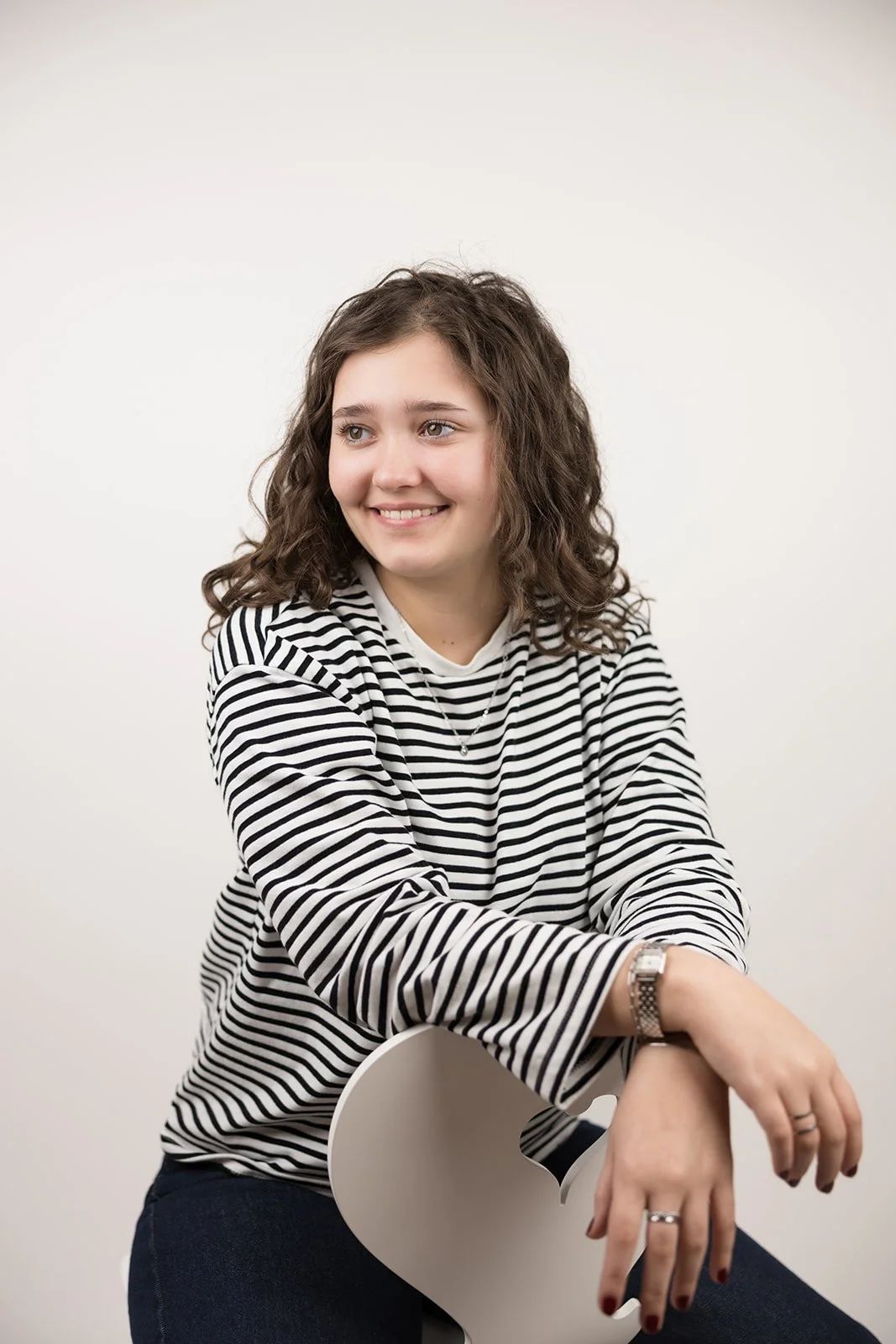 Junge Frau mit lockigem braunem Haar, trägt ein schwarz-weiß gestreiftes Oberteil und sitzt auf einem weißen Stuhl vor weißem Hintergrund, lächelnd und in entspannter Pose.