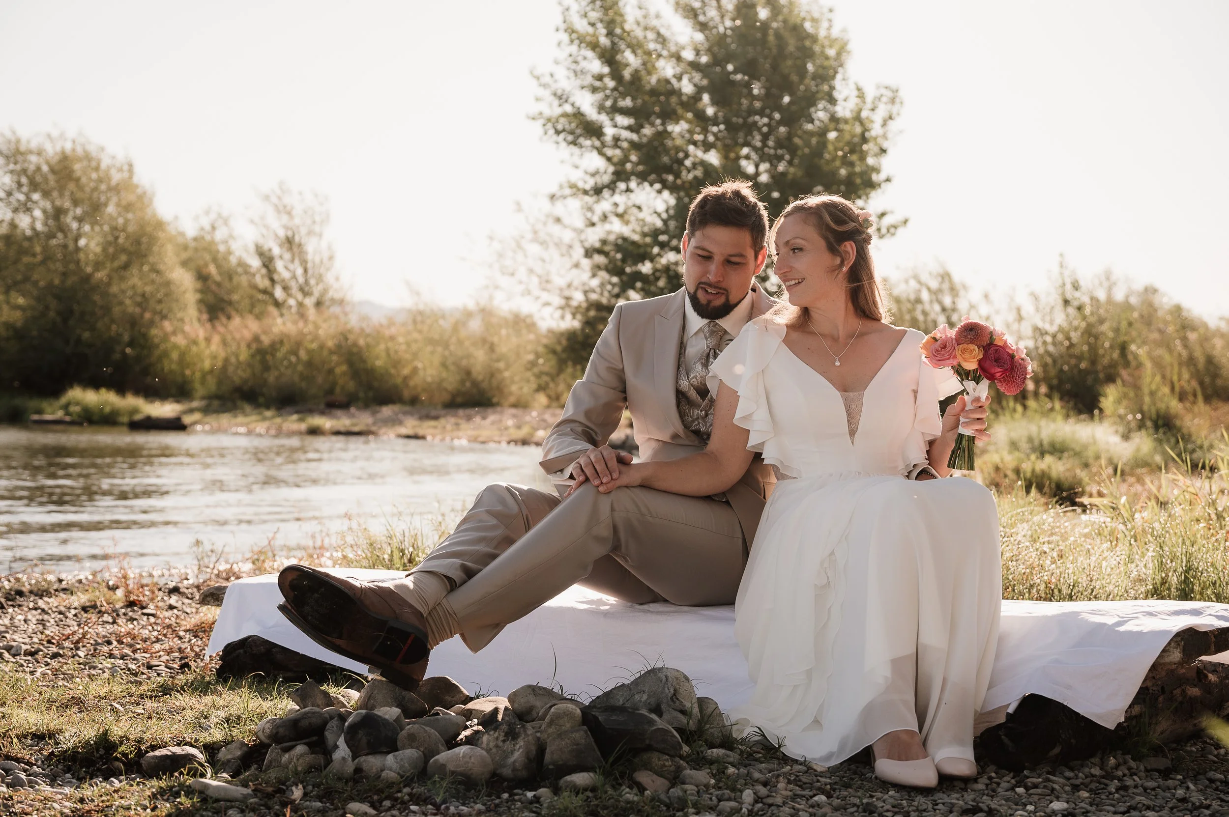 Ein Paar sitzt an einem Flussufer, Frau in einem weißen Kleid mit Blumenbund, er in einem Anzug, bei Sonnenuntergang.