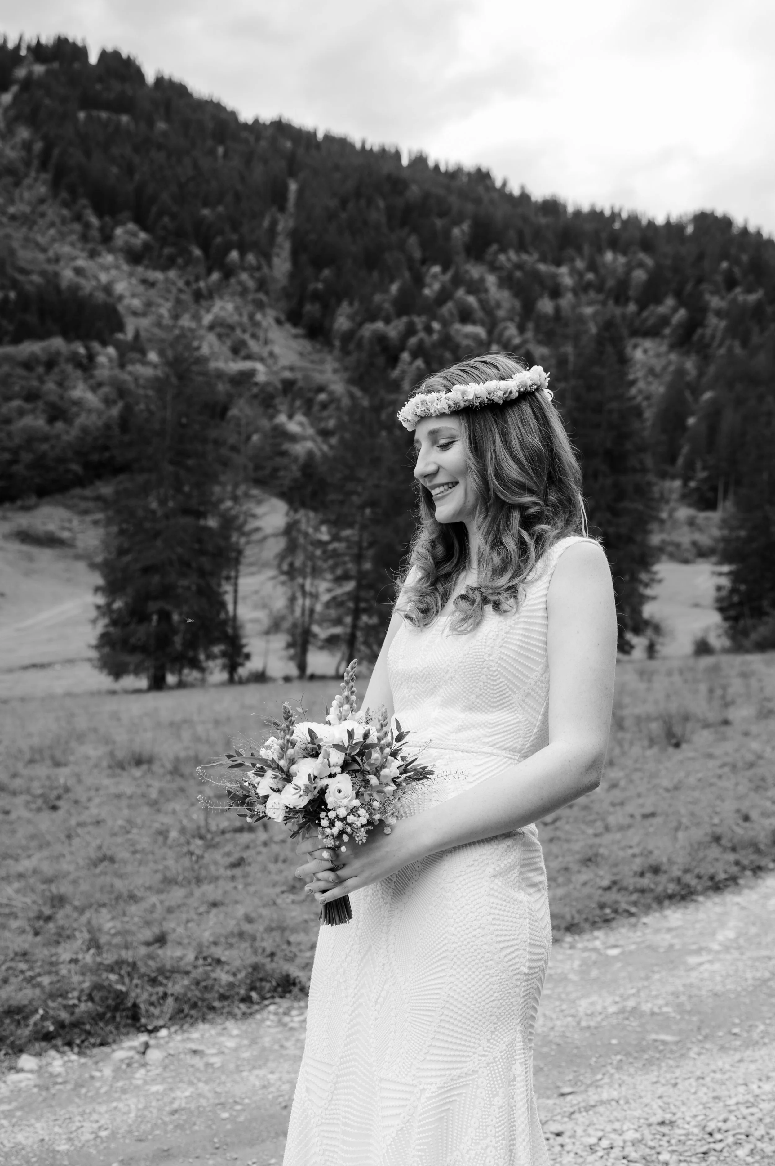 Eine lachende Frau in einem weißen Kleid mit Blumenkranz auf dem Kopf hält einen Blumenstrauß in einem malerischen, bewaldeten Berggebiet.