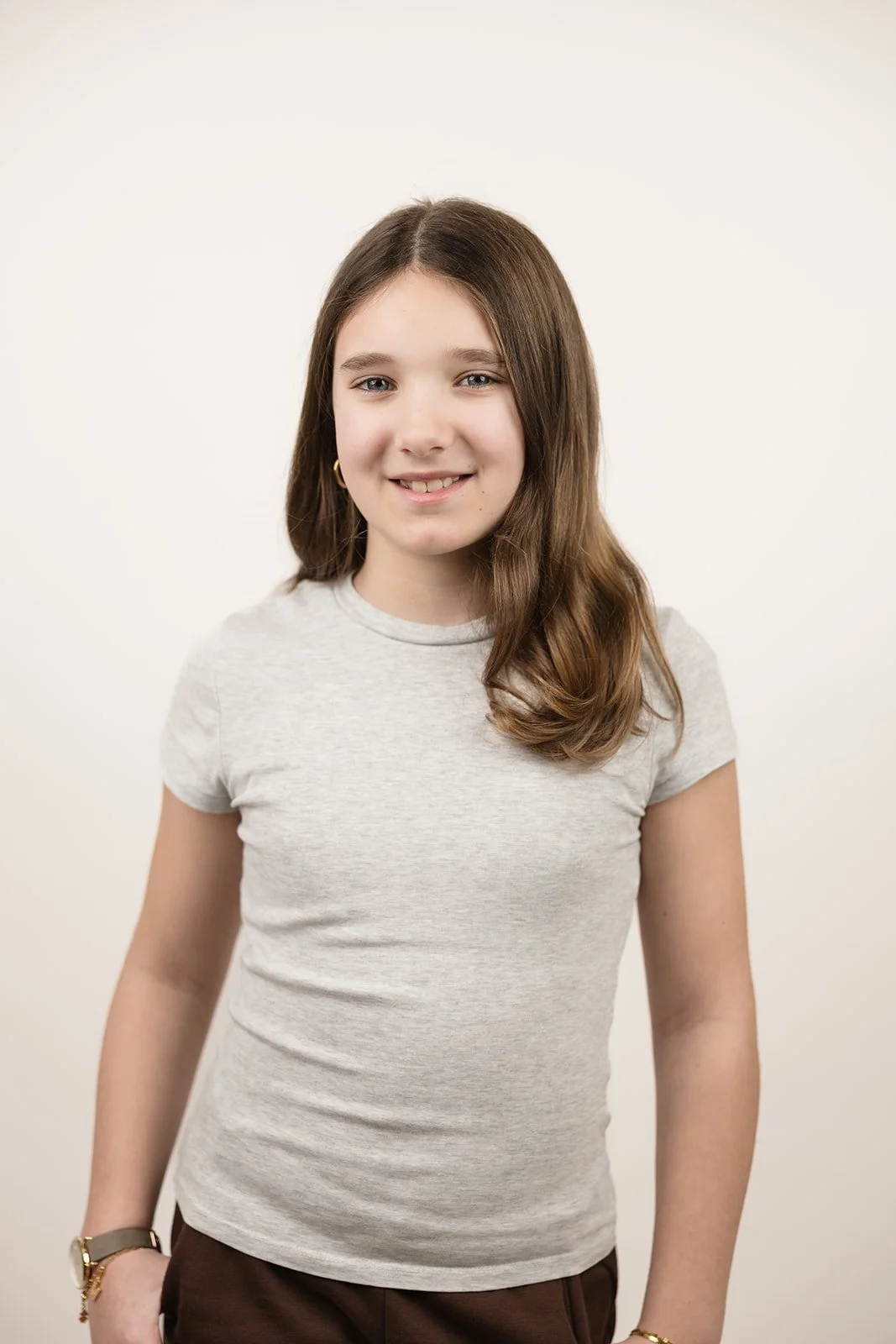 Ein junges Mädchen mit braunen Haaren, hellgrauem T-Shirt und braunen Hosen, das vor einem weißen Hintergrund steht und lächelt.