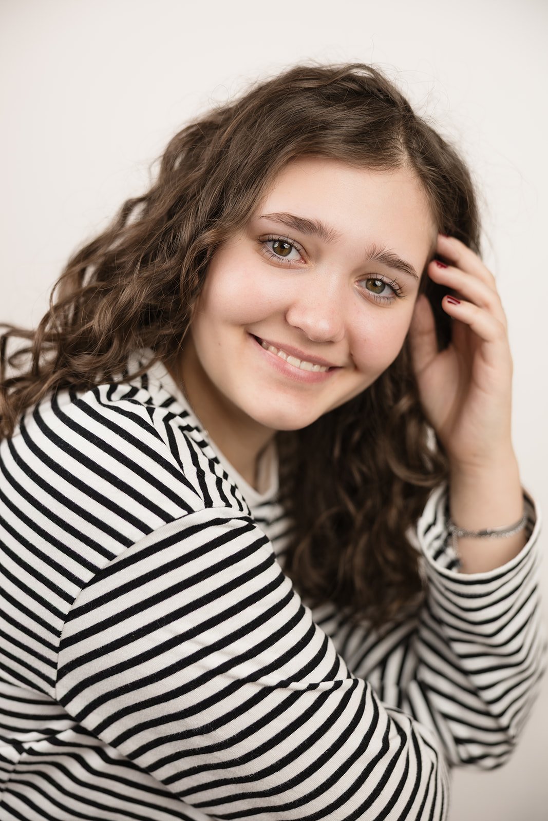 Ein junges Mädchen mit braunen, lockigen Haaren, die lächelnd in die Kamera schaut, trägt ein schwarz-weiß gestreiftes Hemd.