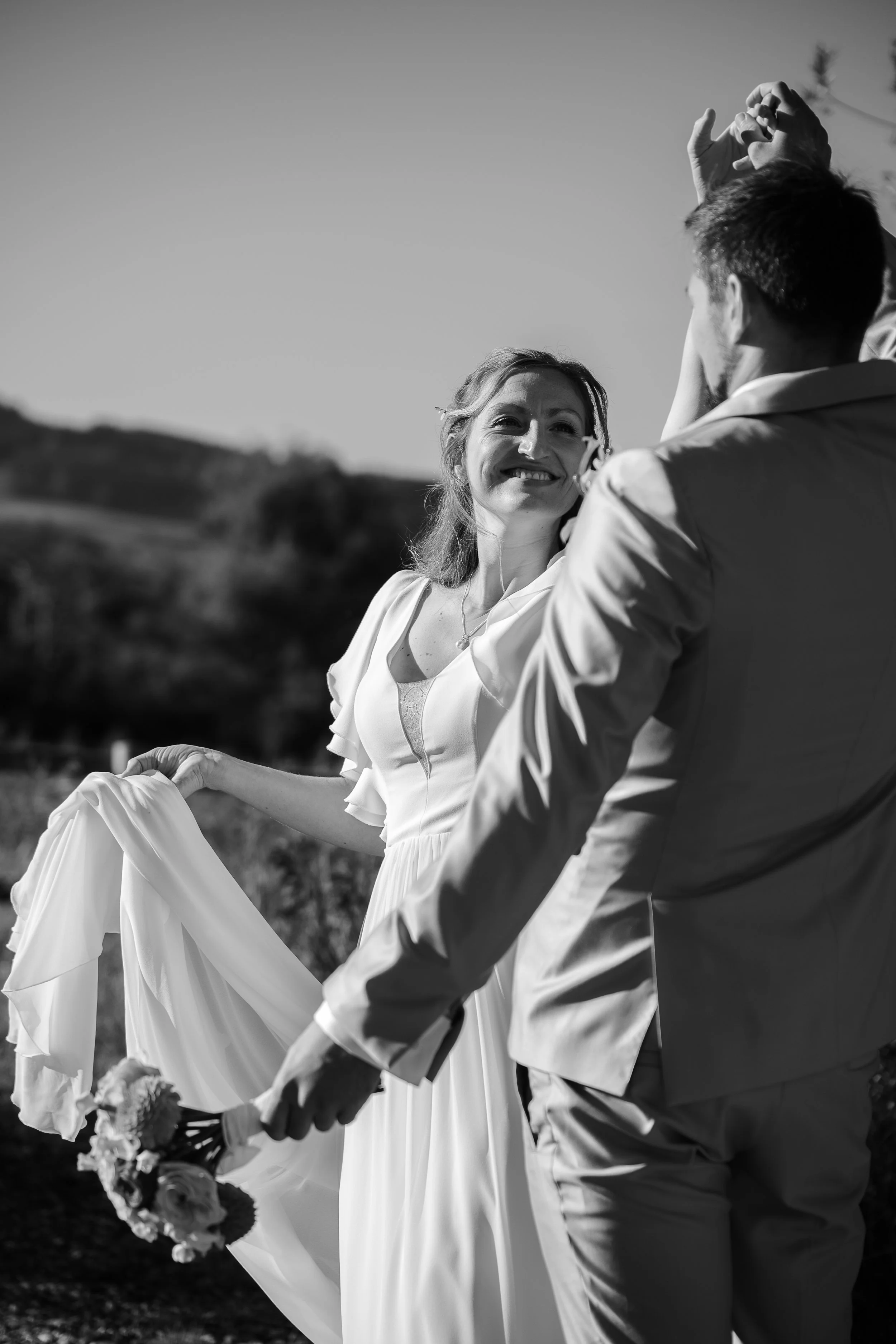 Braut und Bräutigam tanzen im Freien bei Sonnenuntergang, sie lächeln sich an, die Braut trägt ein langes weißes Kleid, der Bräutigam einen Anzug, im Hintergrund sind Hügel und Bäume sichtbar.