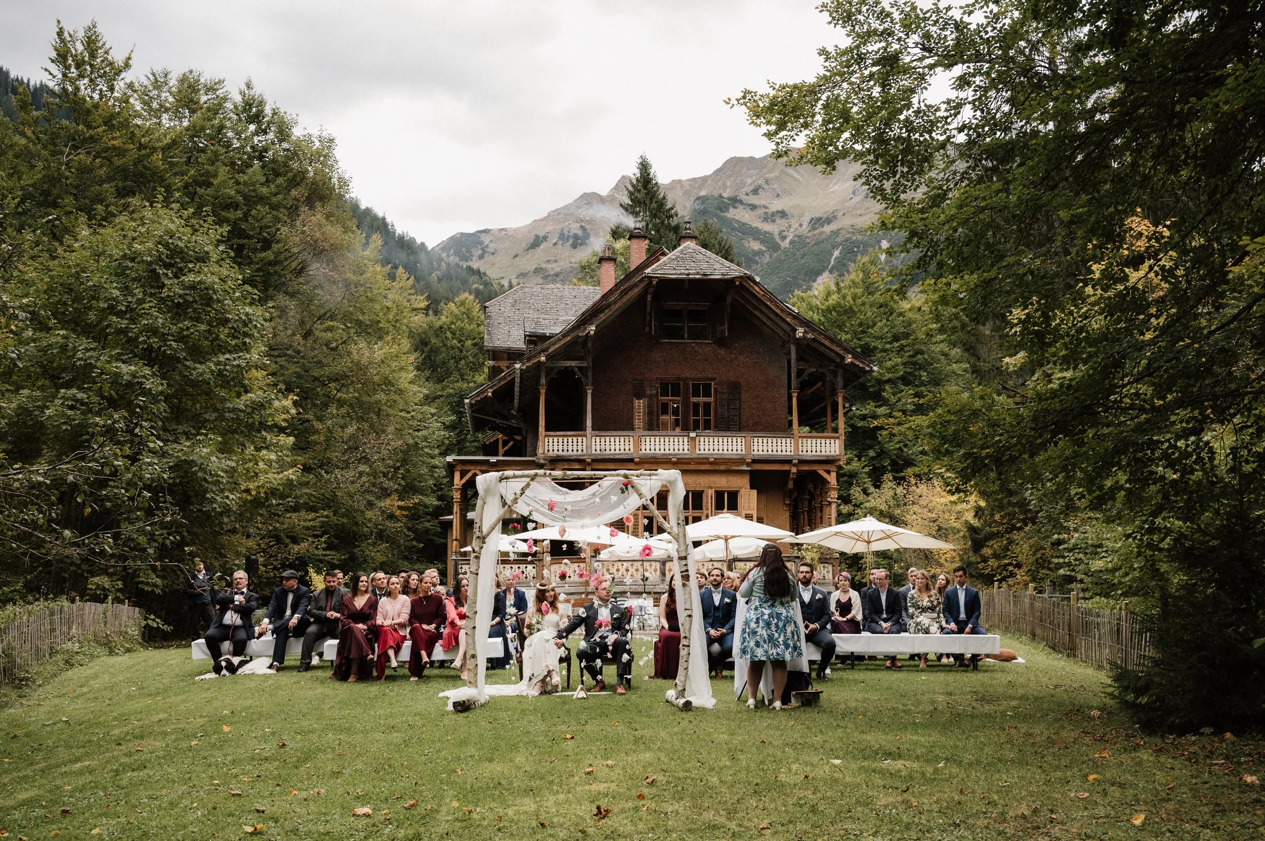 Hochzeit im Grünen vor einem großen Holzhaus mit einem Arkaufbau im Vordergrund.