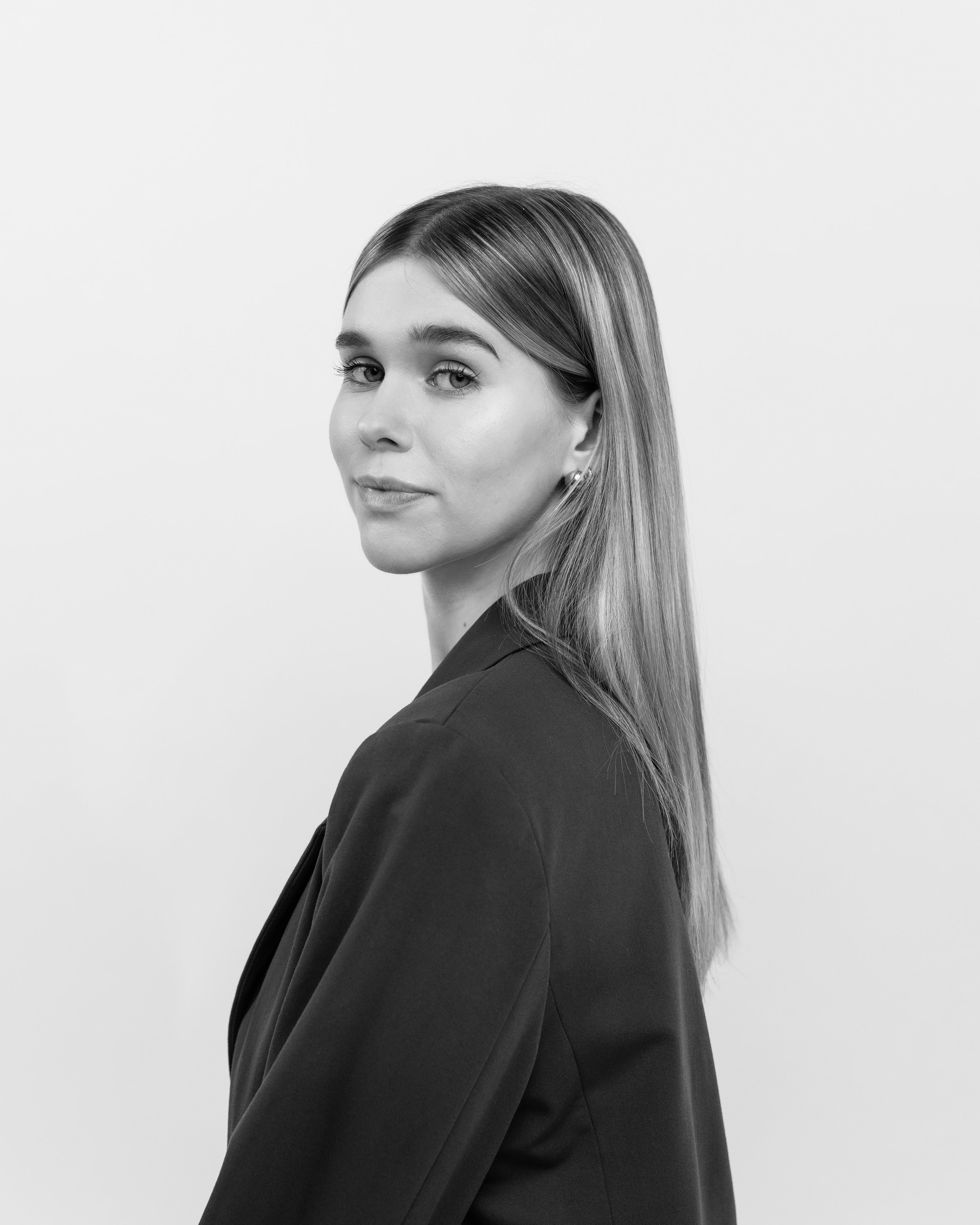 Schwarz-weiß Porträt einer jungen Frau mit langem Haar, getragen in einem dunklen Anzug, blickt über die Schulter, neutraler Ausdruck, helles, schlichtes Hintergrund.