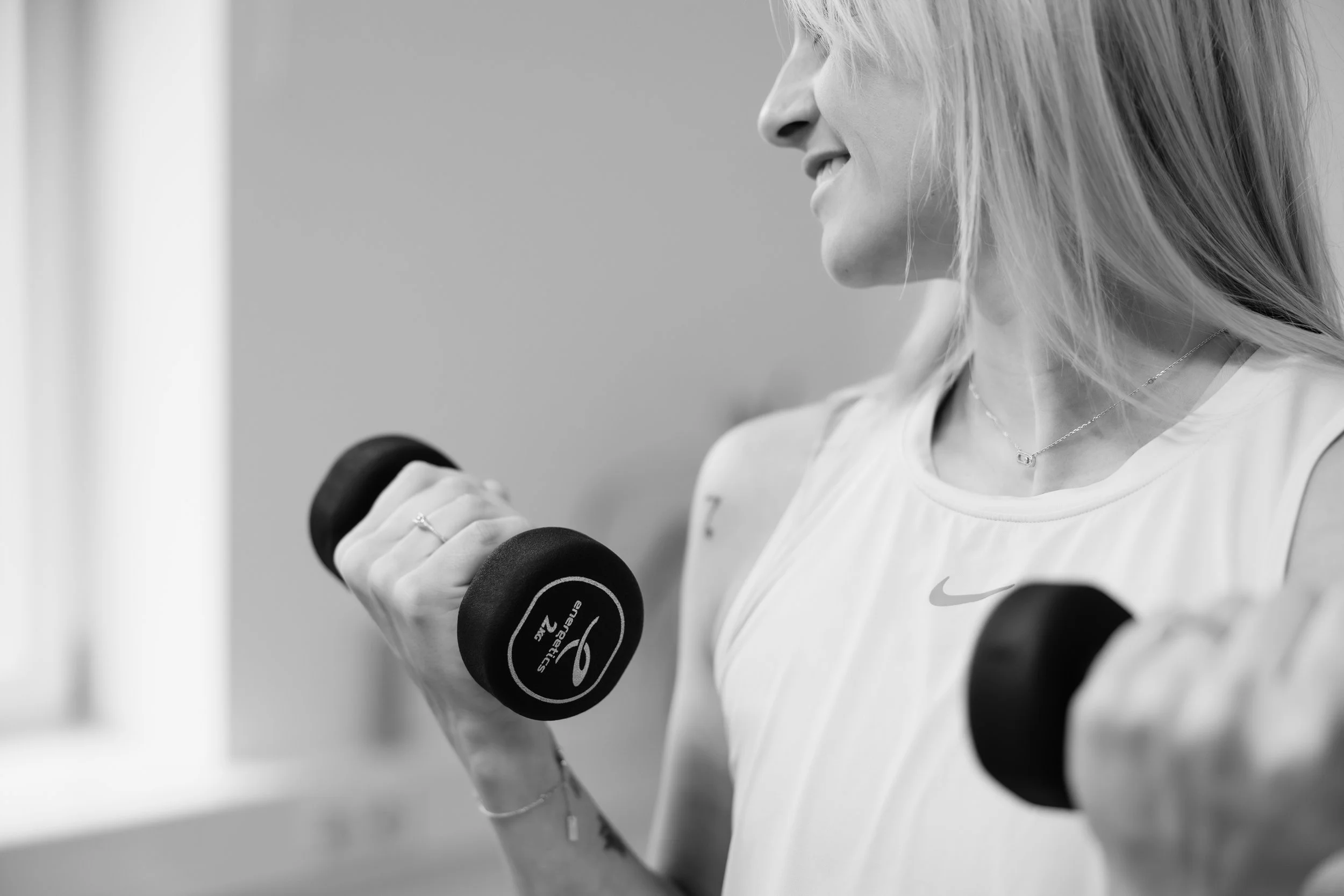 Frau trainiert mit Hanteln in einem Fitnessstudio, lachend und aufrecht stehend, sportliche Kleidung, Cappuccino in sportlicher Kleidung, schwarz-weiß Foto.