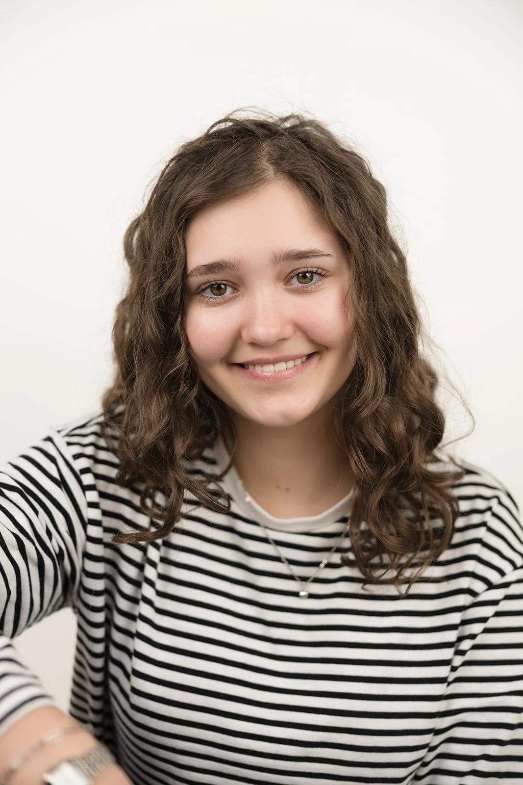 Junge Frau mit braunen, lockigen Haaren und einem gestreiften schwarzen und weißen Hemd, lächelnd vor weißem Hintergrund.
