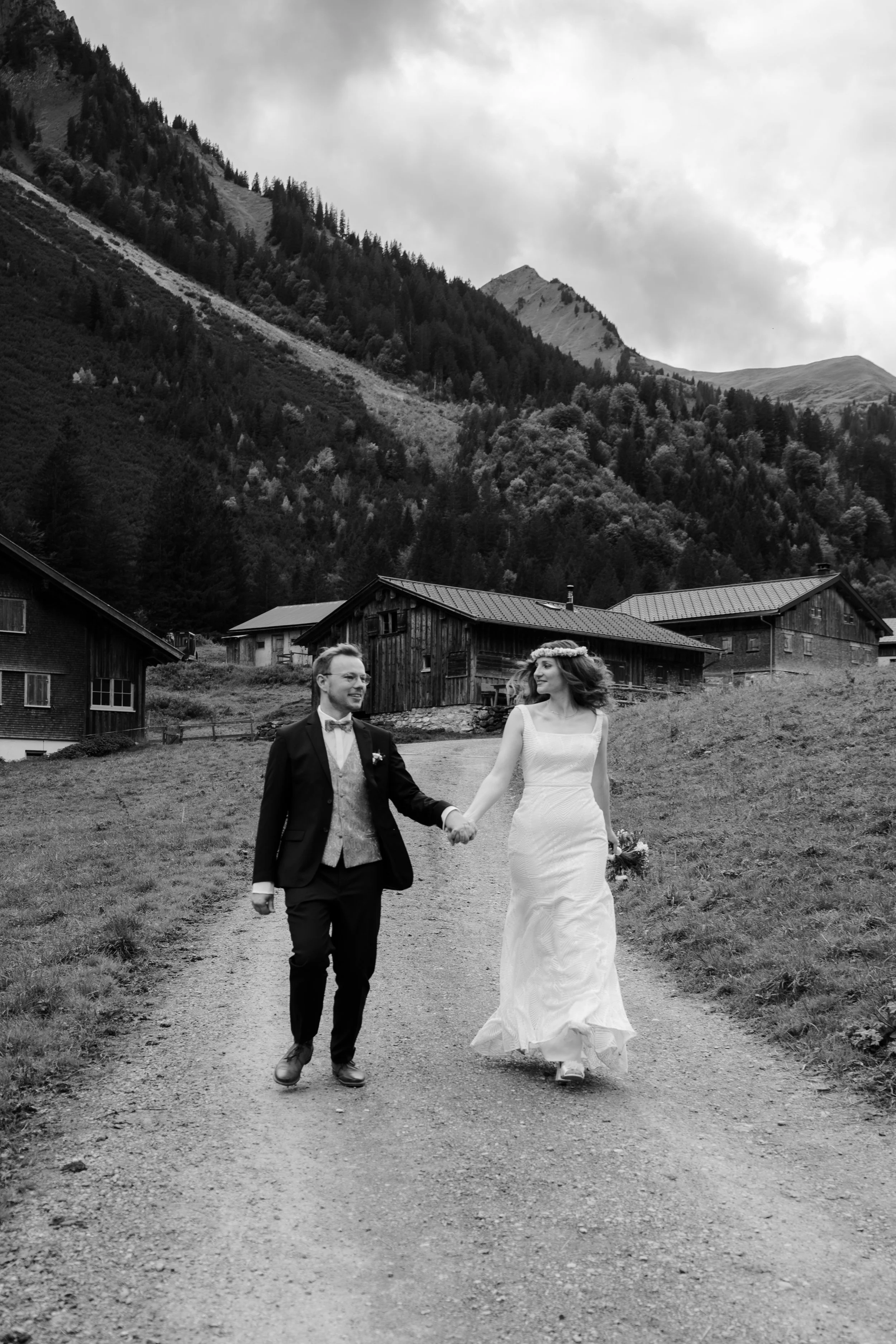 Ein Brautpaar in Hochzeitskleidung, das Hand in Hand auf einem ländlichen Weg zwischen Holzhäusern und Bergen wandert.