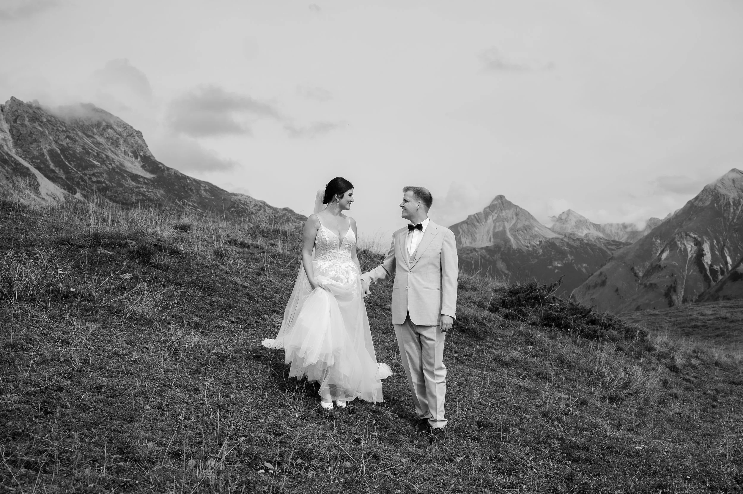 Ein Brautpaar in Hochzeitskleidung steht in einer Berglandschaft, lächelnd und Händchen haltend, in Schwarz-Weiß-Fotografie.