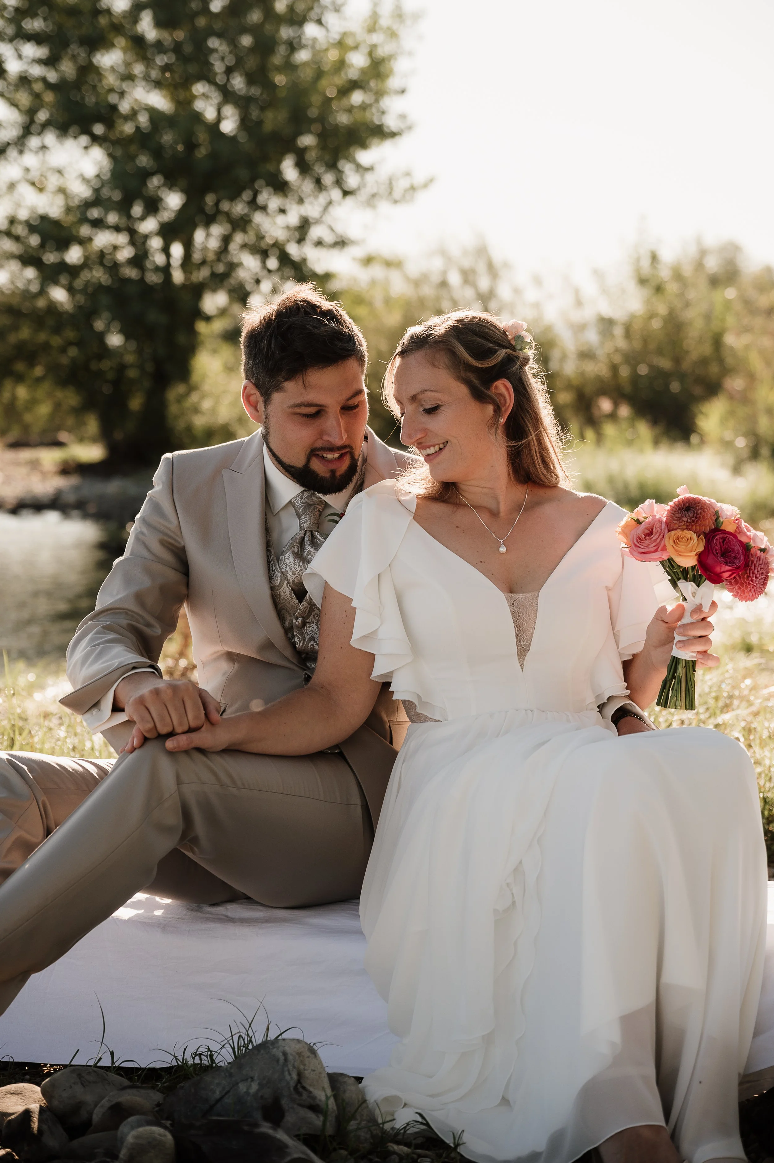 Ein Paar im Hochzeitsoutfit sitzt draußen an einem Fluss, umgeben von Natur, lächelnd und sich verbunden.