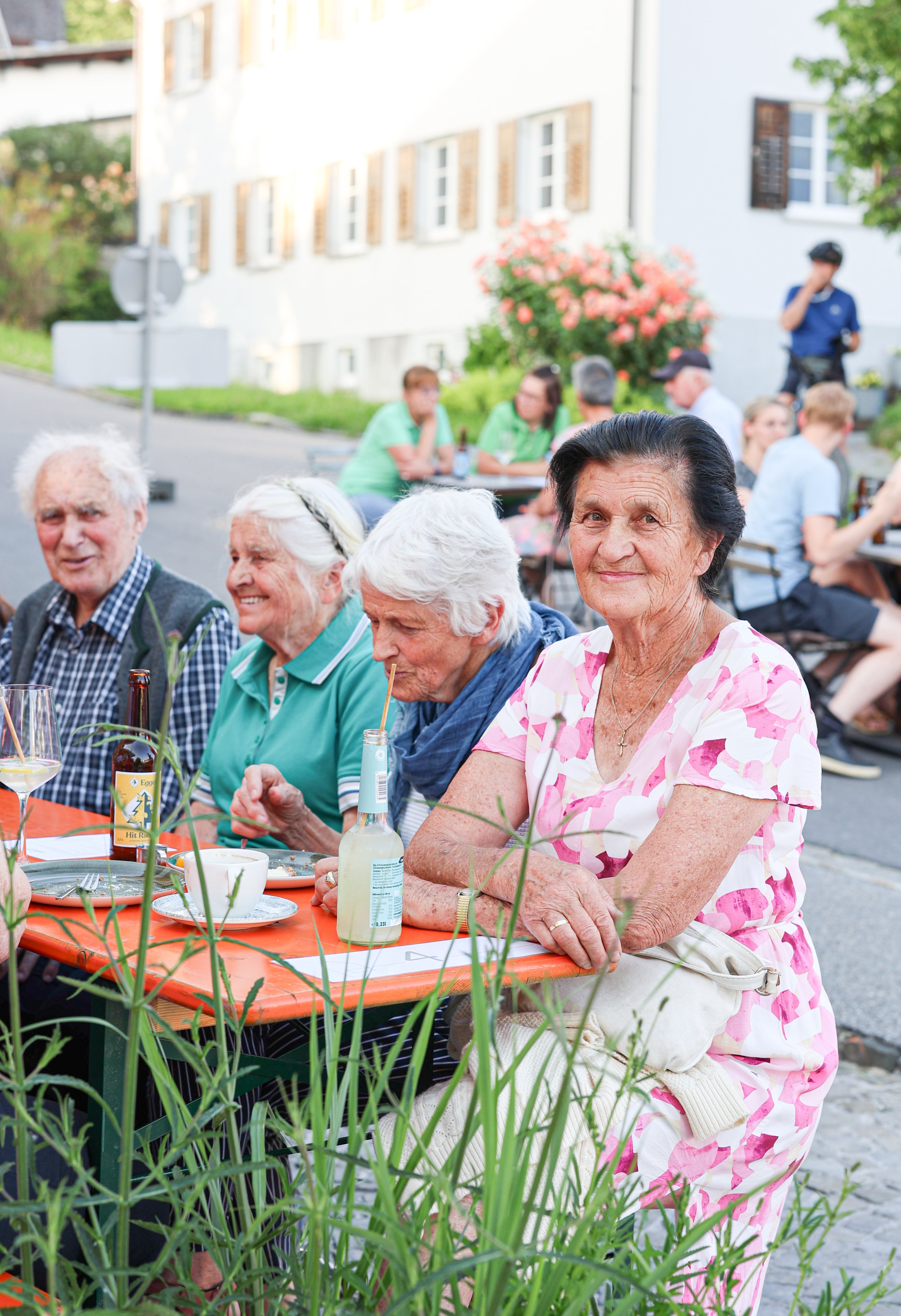 Eine Gruppe älterer Menschen sitzt bei einer Veranstaltung im Freien, einige trinken und essen, im Hintergrund sind weitere Personen, ein Gebäude und eine blühende Pflanze zu sehen.