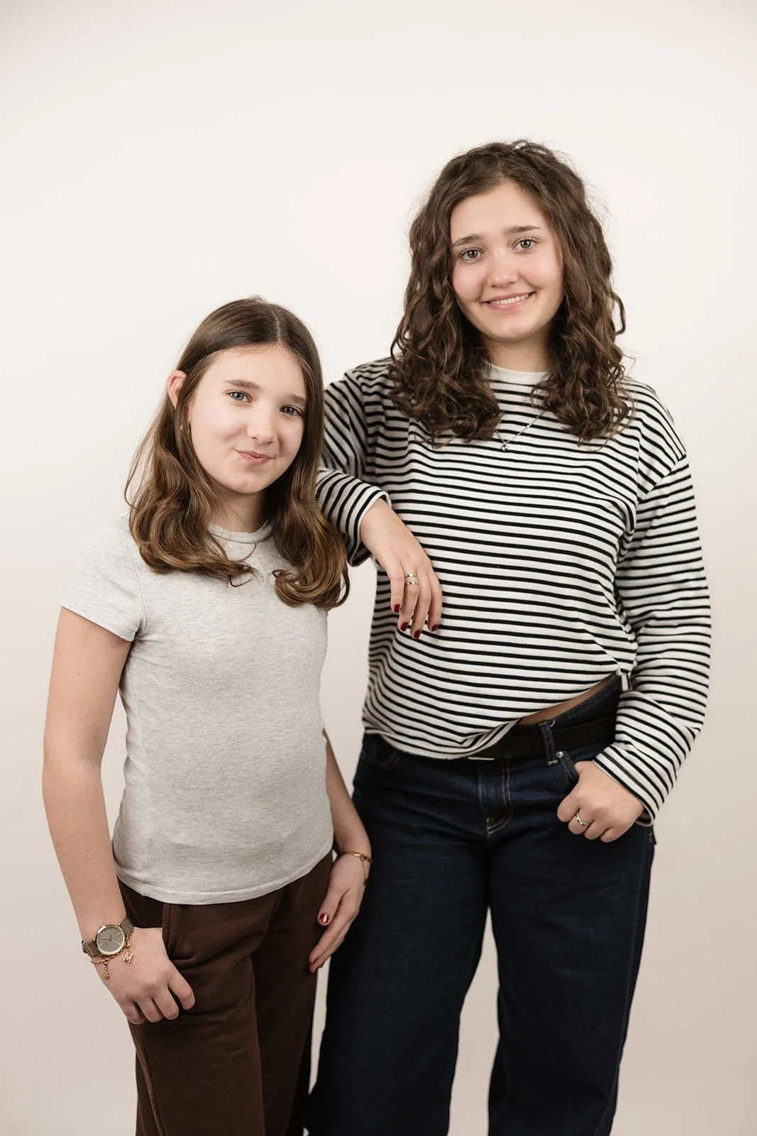 Zwei junge Frauen vor weißem Hintergrund, lächelnd, casual Kleidung, eine mit gestreiftem T-Shirt, die andere mit grauem T-Shirt und braunen Hosen.