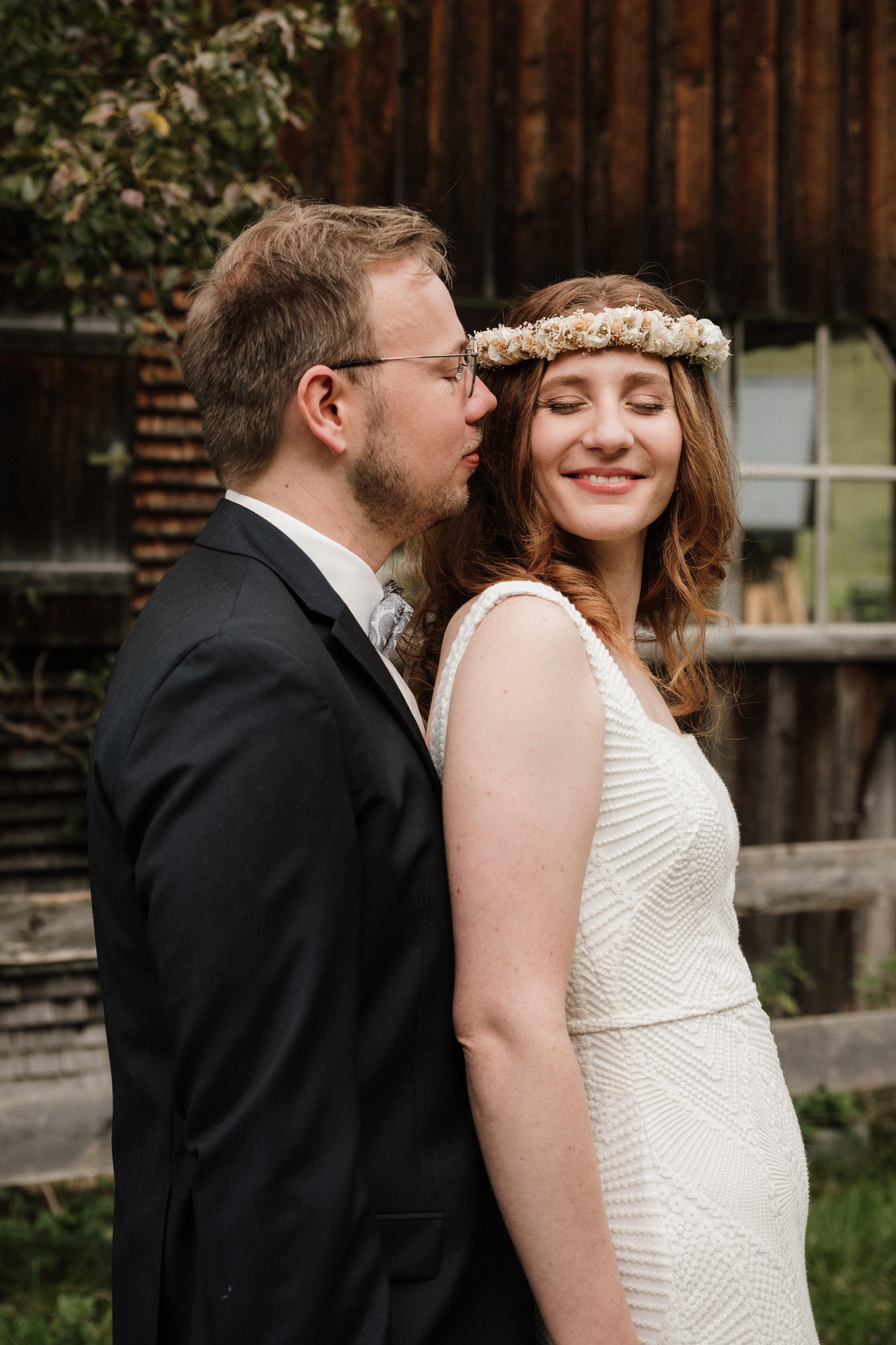 Ein Brautpaar, ein Mann in einem schwarzen Anzug und eine Frau in einem weißen Kleid mit einem Blumenkranz im Haar, steht draußen vor einem Holzgebäude.