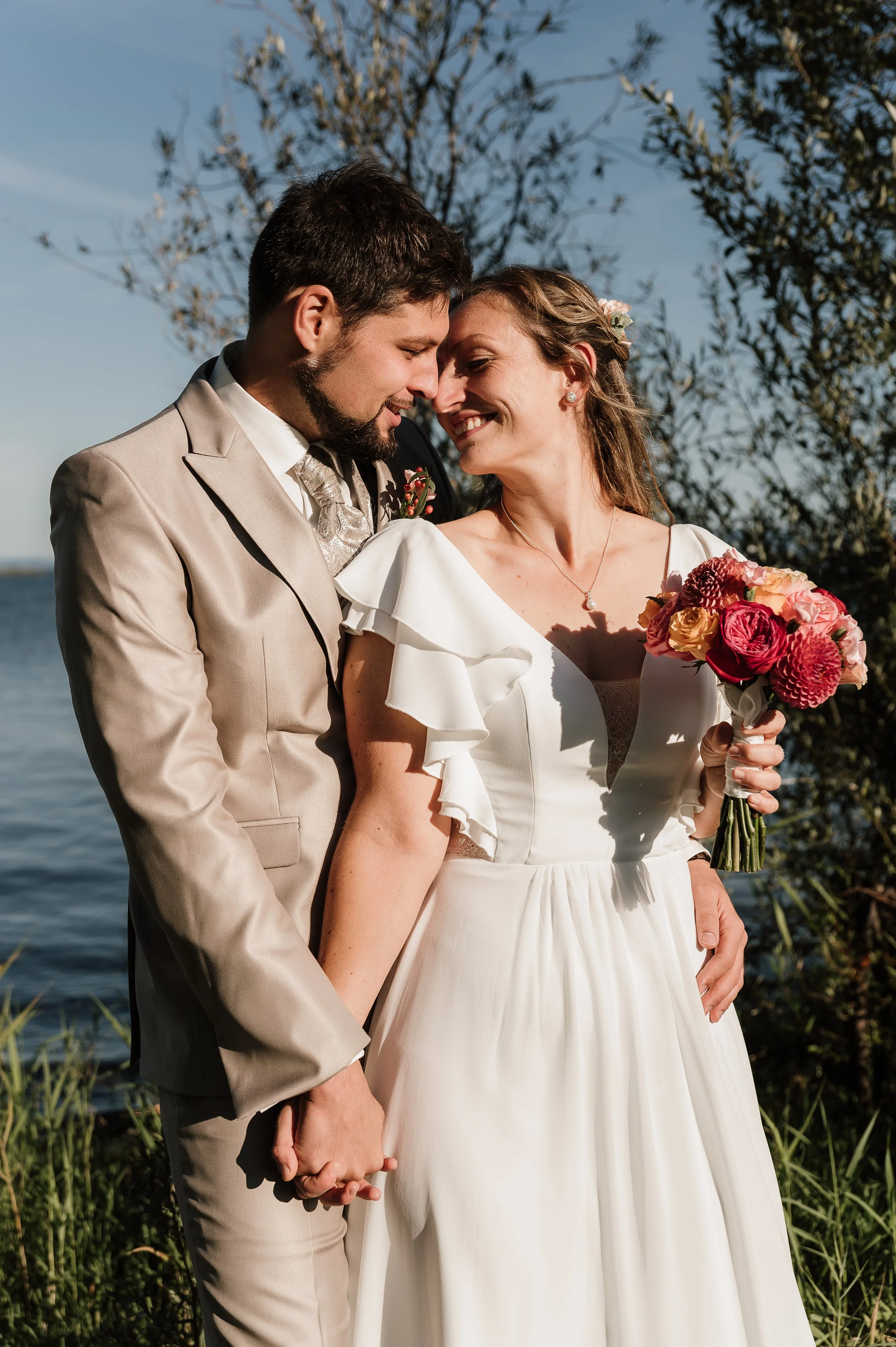Ein Brautpaar bei einer Hochzeit, die Hand in Hand stehen, nah beieinander, vor einem Wasserhintergrund und Bäumen bei Sonnenuntergang.