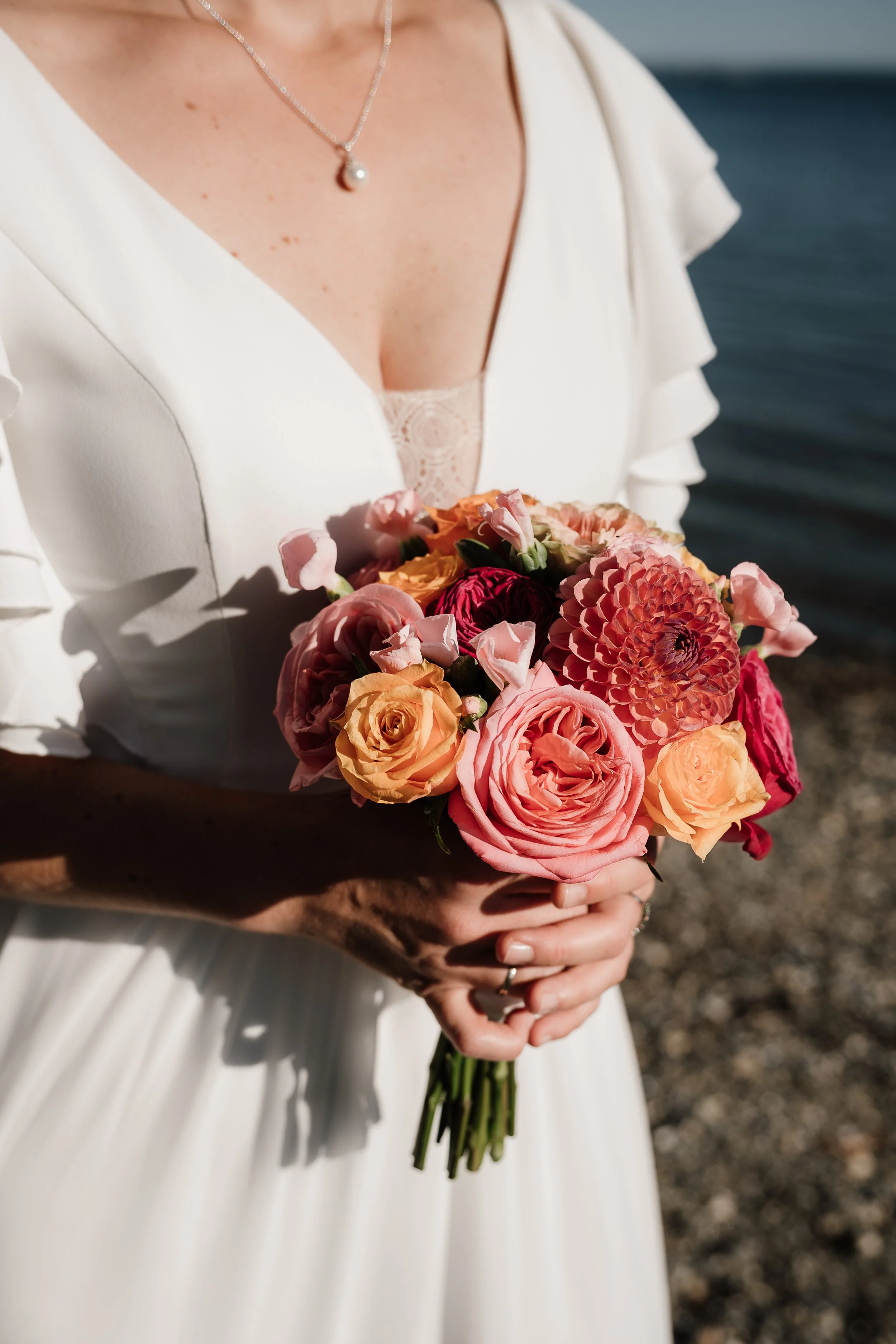 Braut hält einen bunten Blumenstrauß bei Sonnenuntergang am Meer.