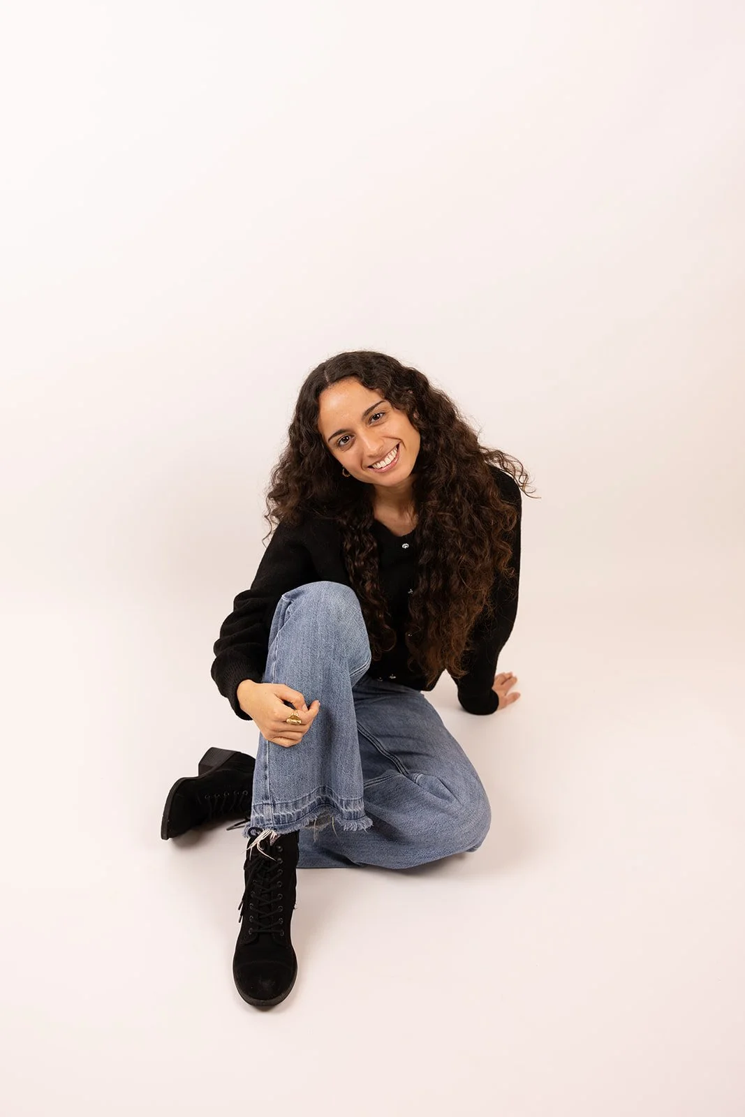 Junge Frau mit langen, lockigen Haaren, lachend, sitzt auf dem Boden mit einem Bein angezogen, trägt schwarze Stiefel, blaue Jeans und schwarze Jacke, vor weißem Hintergrund.