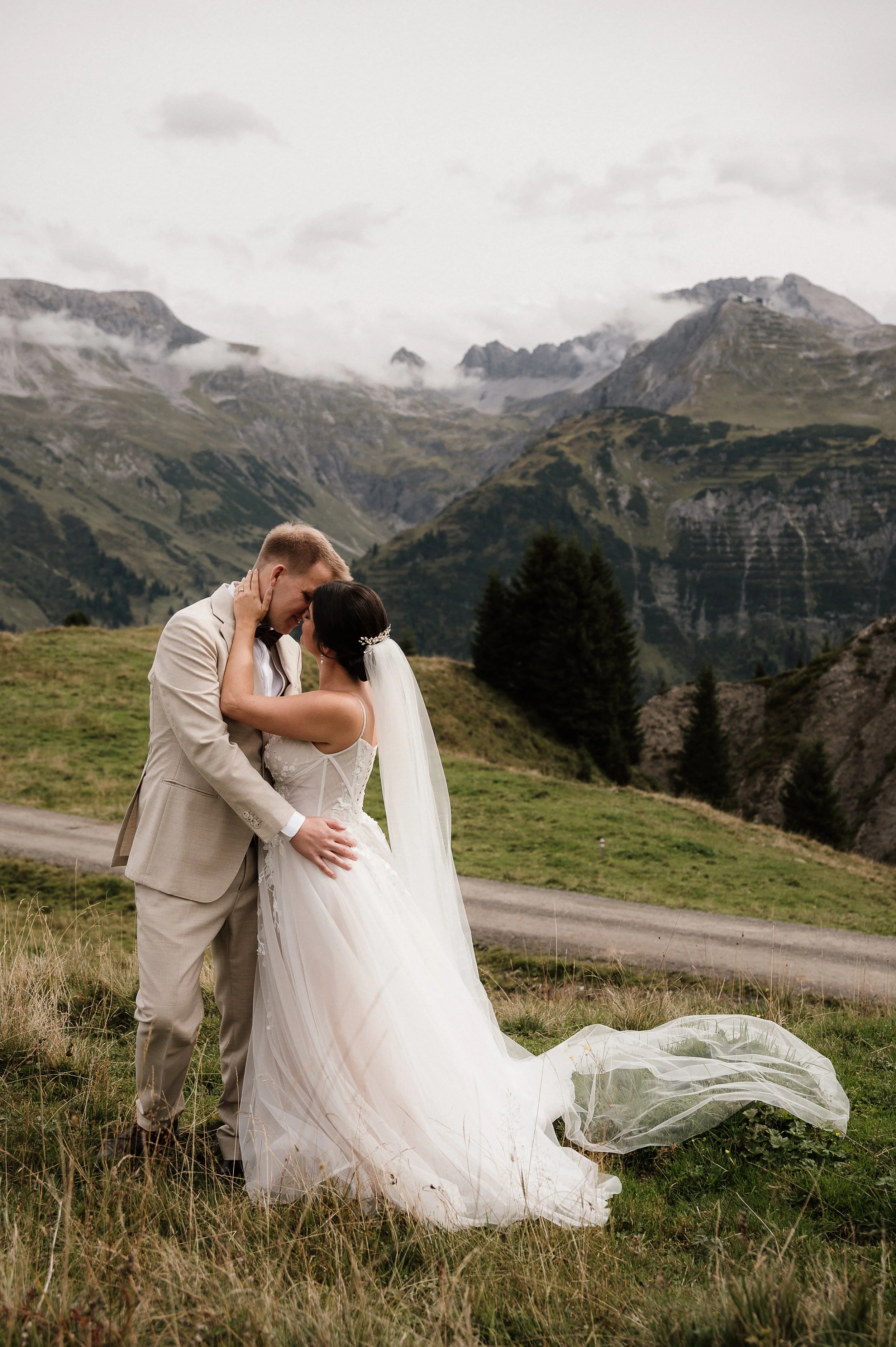 Ein Brautpaar steht sich in den Bergen gegenüber, umarmt sich und küsst sich bei ihrer Hochzeit, umgeben von grüner Landschaft und Bergen im Hintergrund.